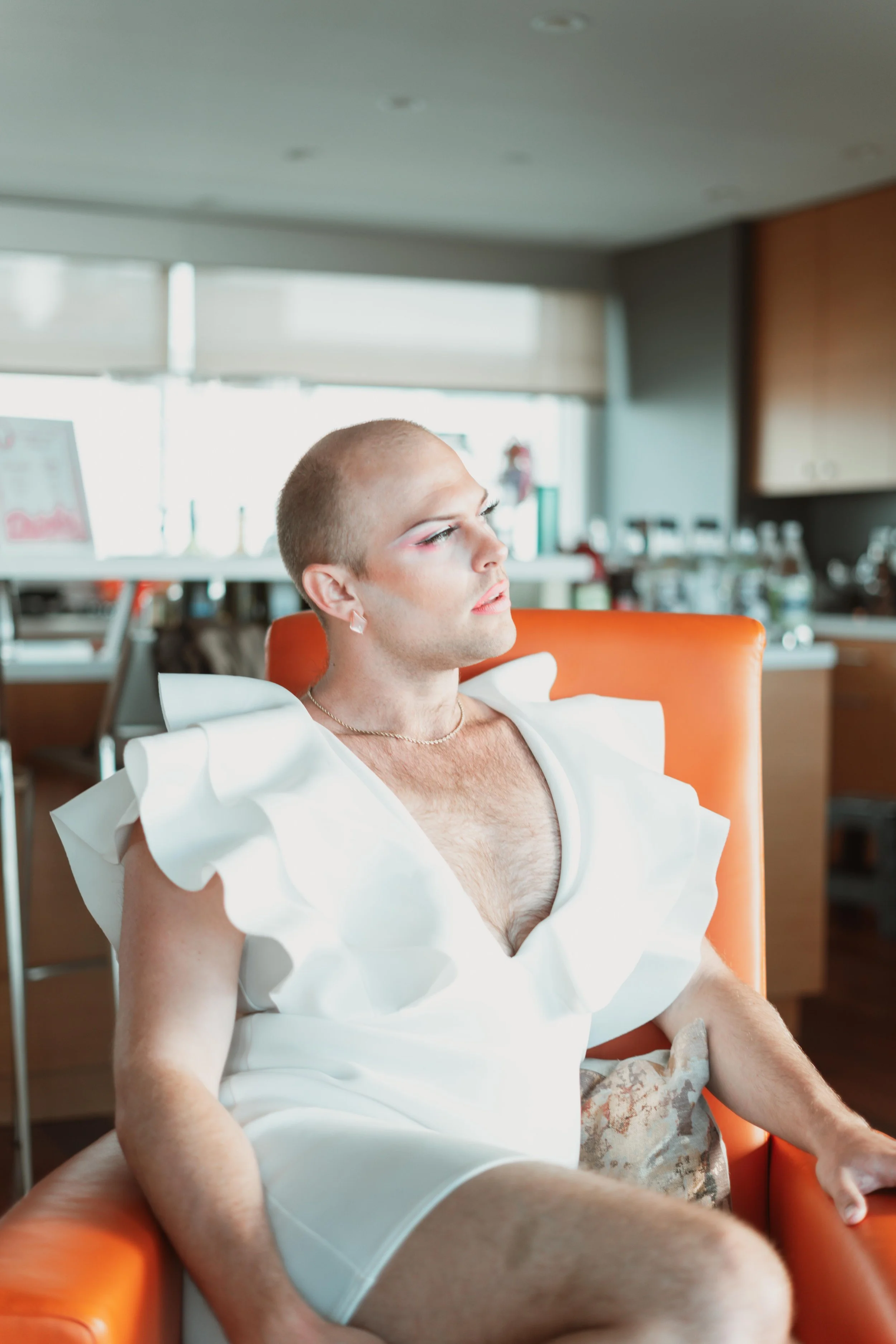A person sitting in an orange chair wearing a white garment with ruffled shoulders, with makeup and jewelry, in a room with natural light and kitchen appliances in the background.