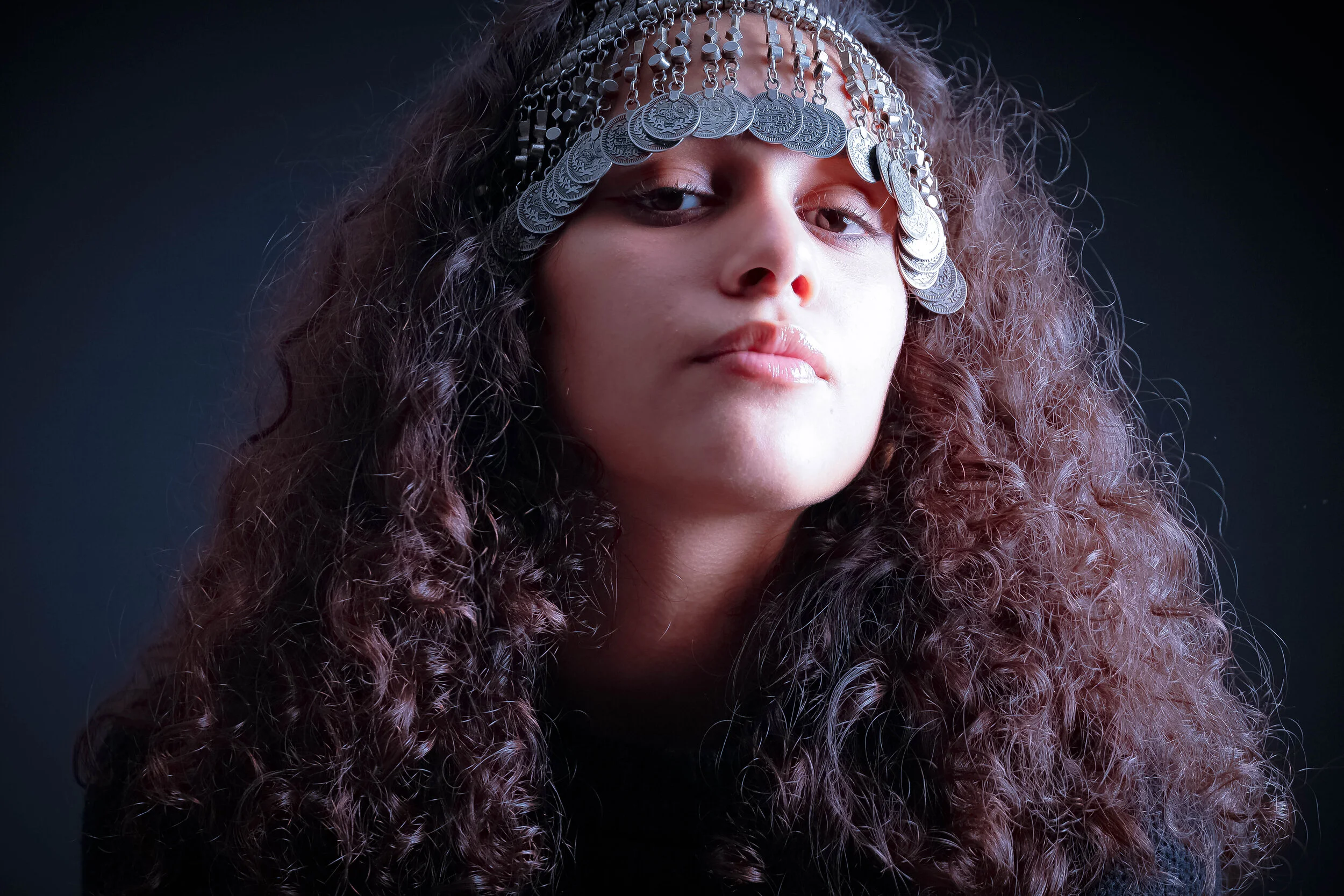 A woman with long, curly hair wearing a headpiece made of hanging coins, looking directly at the camera with a neutral expression against a dark background.