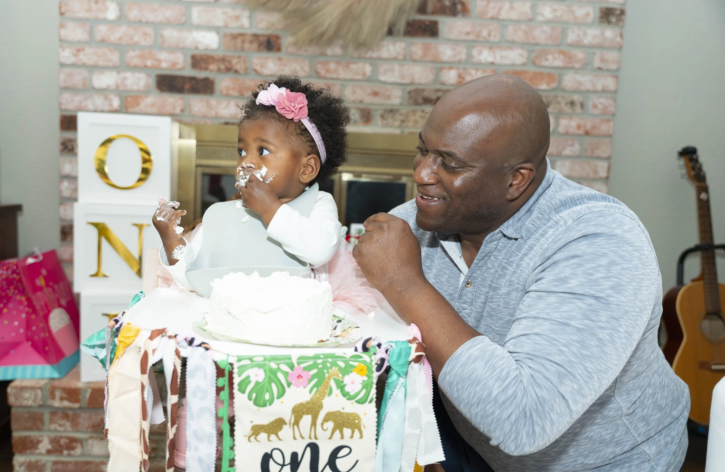 A young girl with a pink headband and white shirt sitting at a birthday cake with white frosting, being celebrated by a smiling man, likely her father, in a living room with a brick fireplace, colorful decorations, and presents.