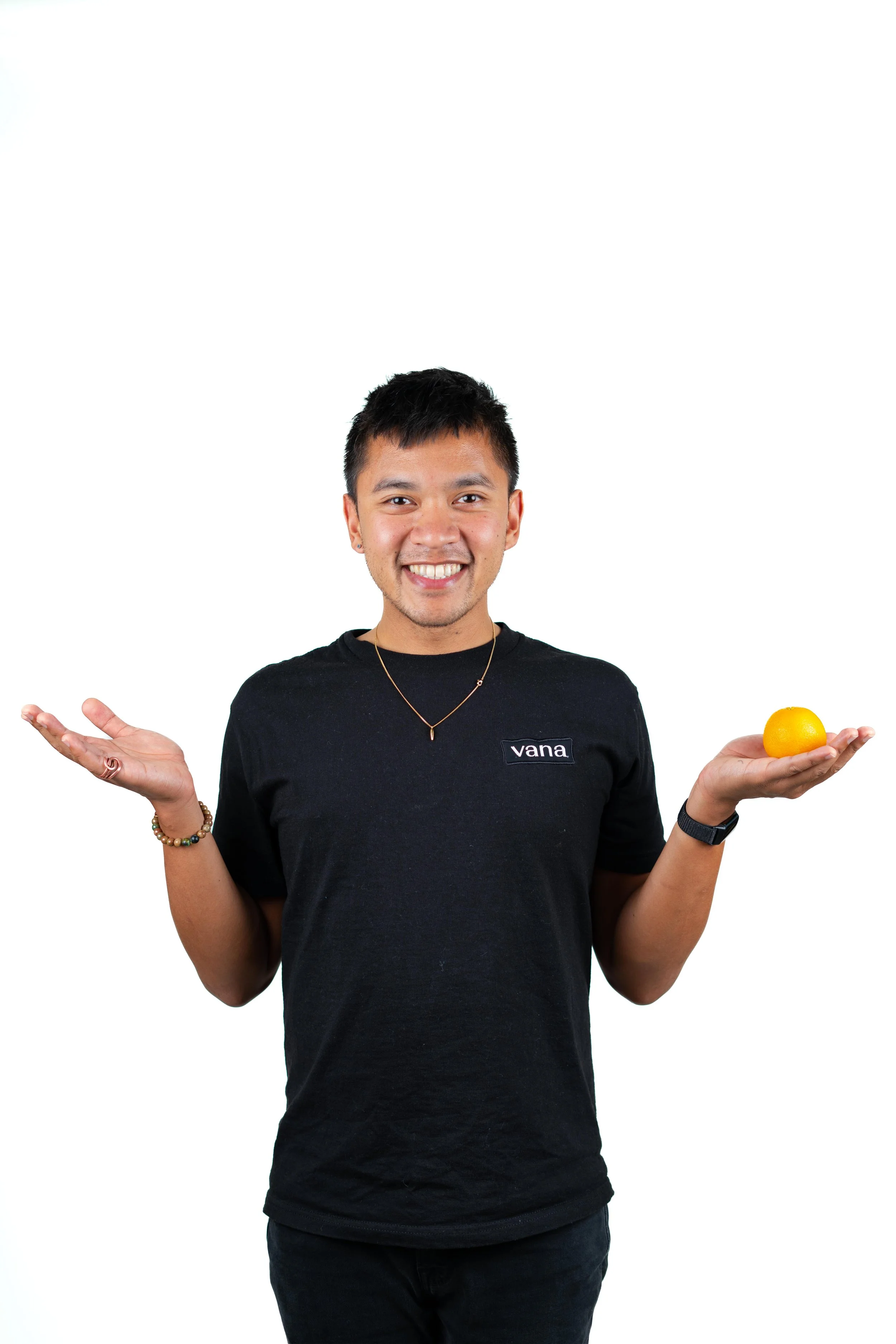 Smiling man holding an orange in his right hand and shrugging with his left hand against a plain white background.