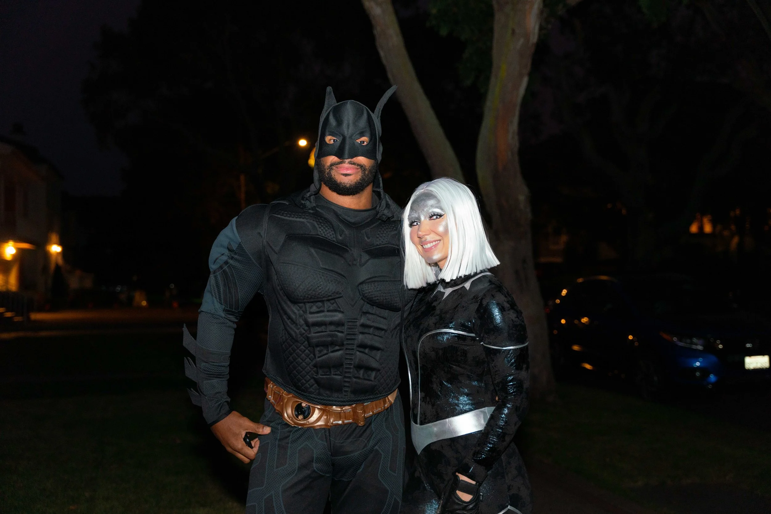 A man dressed as Batman and a woman dressed as a futuristic cyber character pose together outdoors at night.