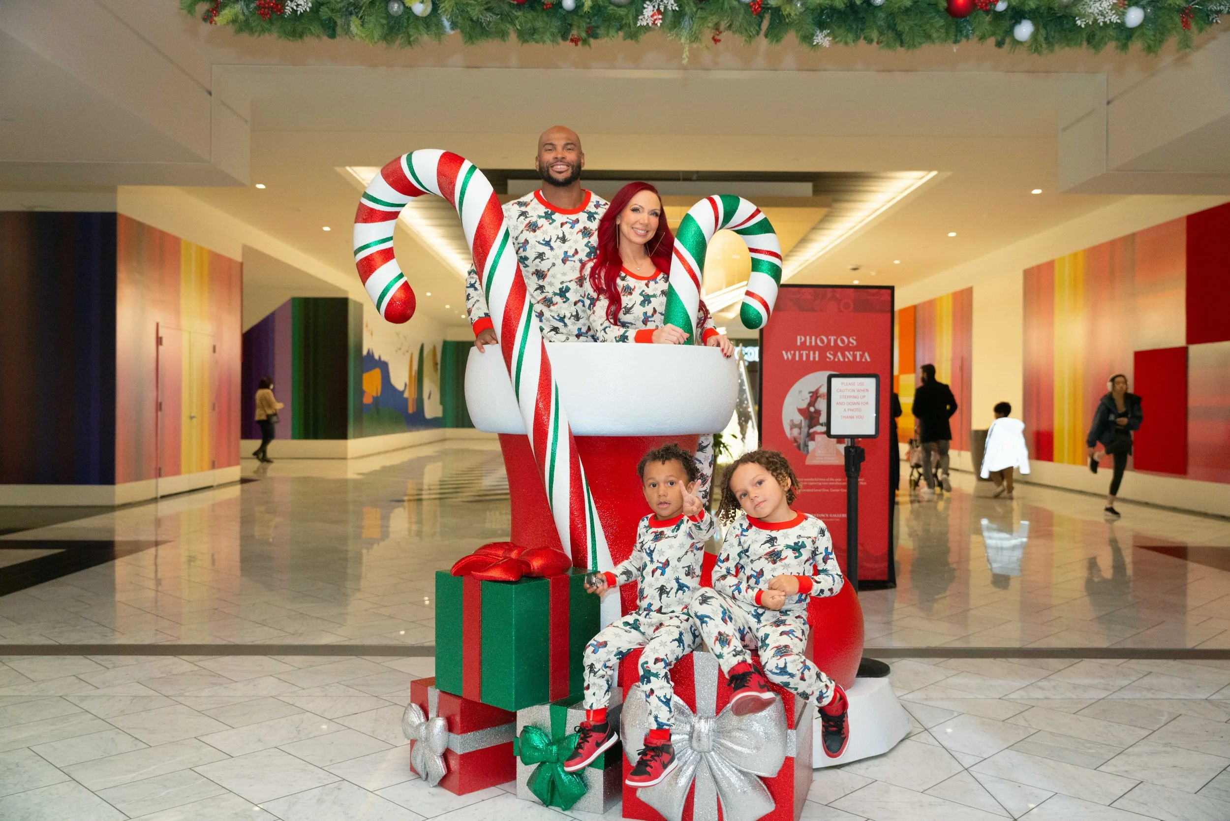 A family of four dressed in matching Christmas pajamas posing with a large holiday display of Santa's boots and candy canes in a mall