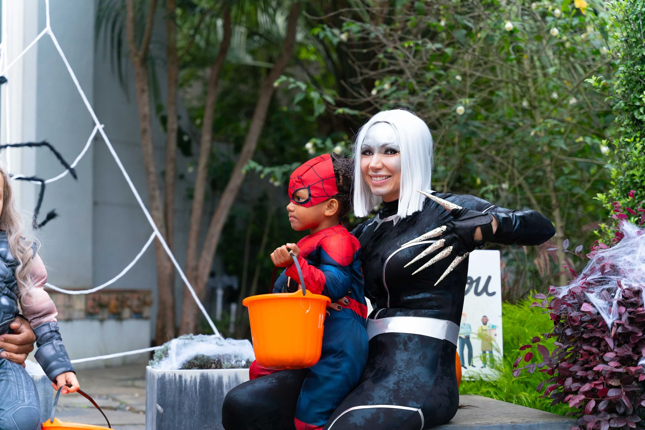 A woman dressed as a dark, spooky character with white hair and black claws, smiling while sitting next to a child in a Spider-Man costume holding a pumpkin bucket, outdoors with Halloween decorations and greenery in the background.