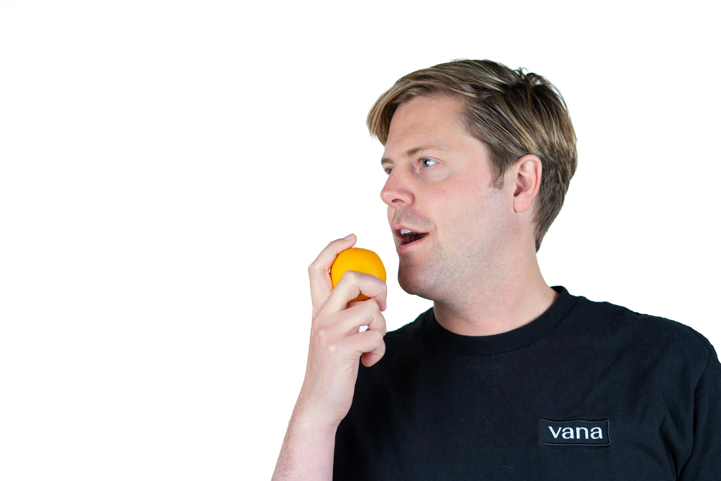 A man with light brown hair, wearing a black T-shirt with a 'vana' logo, holding a lemon near his mouth, appearing to be about to take a bite.