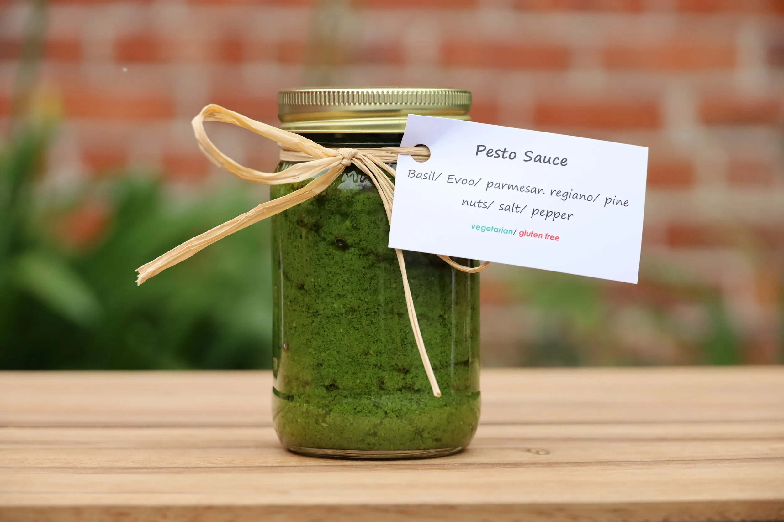A glass jar with a gold lid containing green pesto sauce, tied with a raffia ribbon, with a white label stating 'Pesto Sauce' and ingredients including basil, EVOO, parmesan, regiano, pine nuts, salt, and pepper. The label indicates the pesto is vege