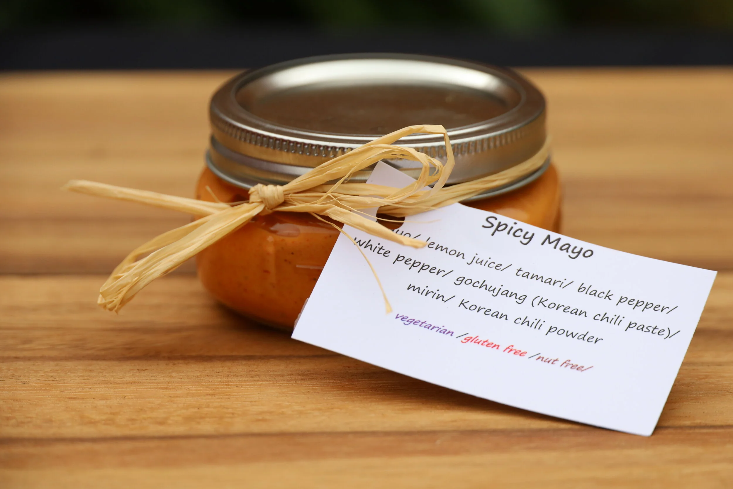 A jar of spicy mayo with a handwritten label tied with a raffia ribbon, sitting on a wooden surface.
