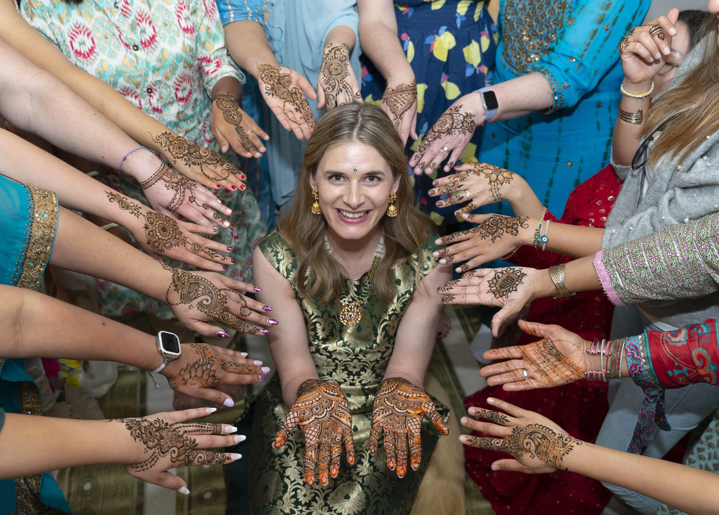 A woman sitting amidst a circle of people, all displaying intricate henna designs on their hands, celebrating an occasion with vibrant traditional attire and jewelry.
