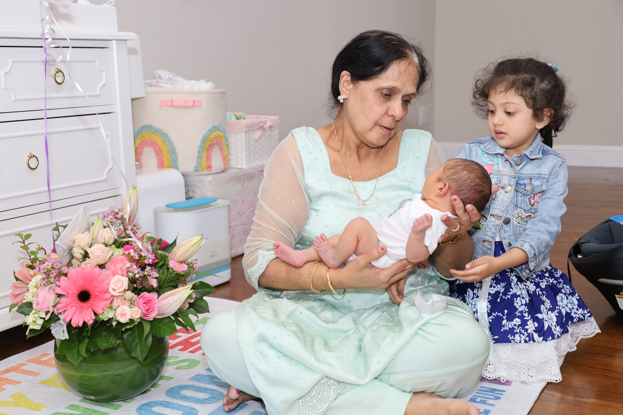 An elderly woman dressed in light green attire is holding a newborn baby while a young girl, wearing a denim jacket and a floral dress, looks on. They are in a room decorated with pastel-colored items and a bouquet of pink flowers.
