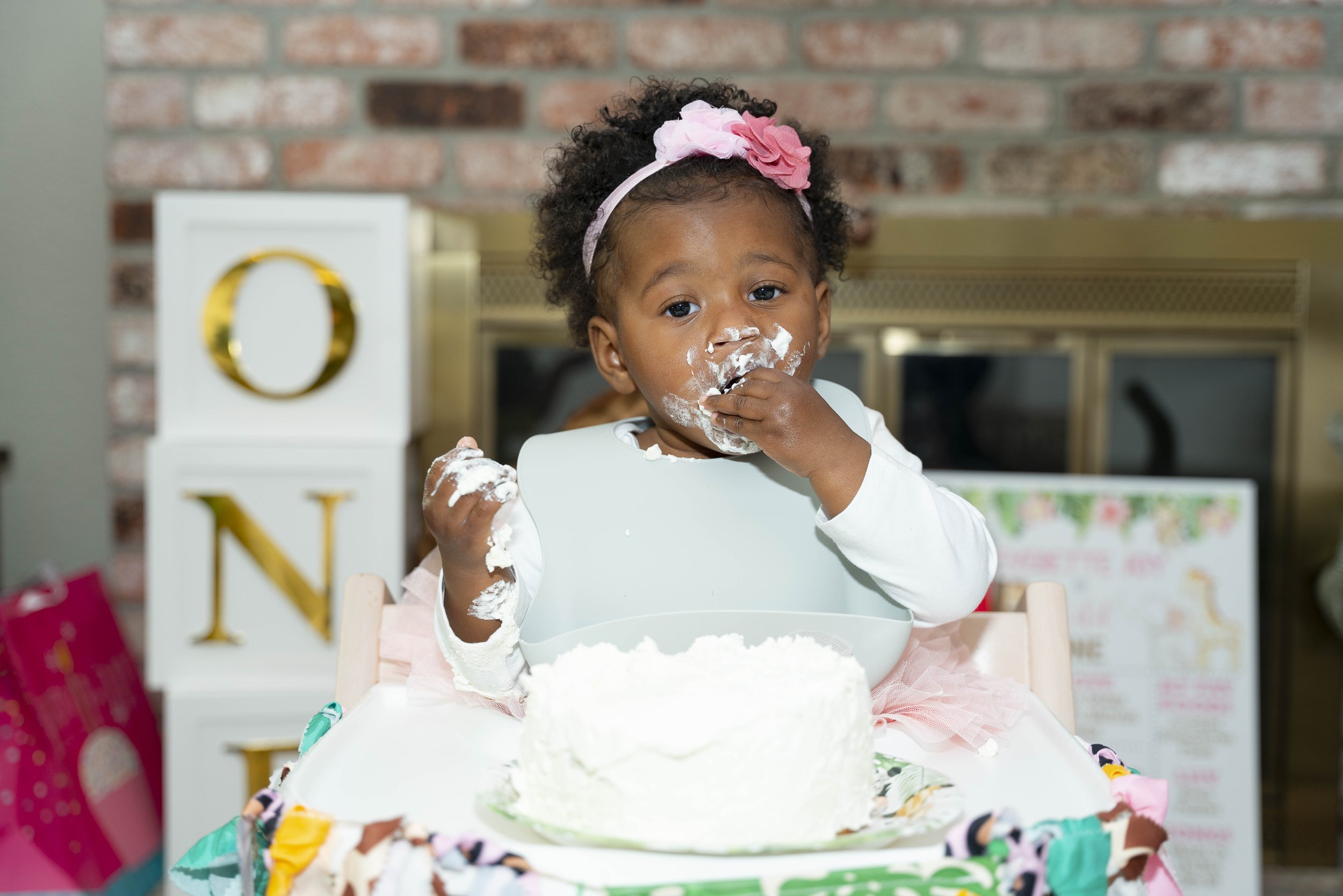 A young girl with curly hair, a pink headband with flowers, eating cake with white frosting at a celebration. She has frosting on her face and hand, sitting in a high chair at a table.