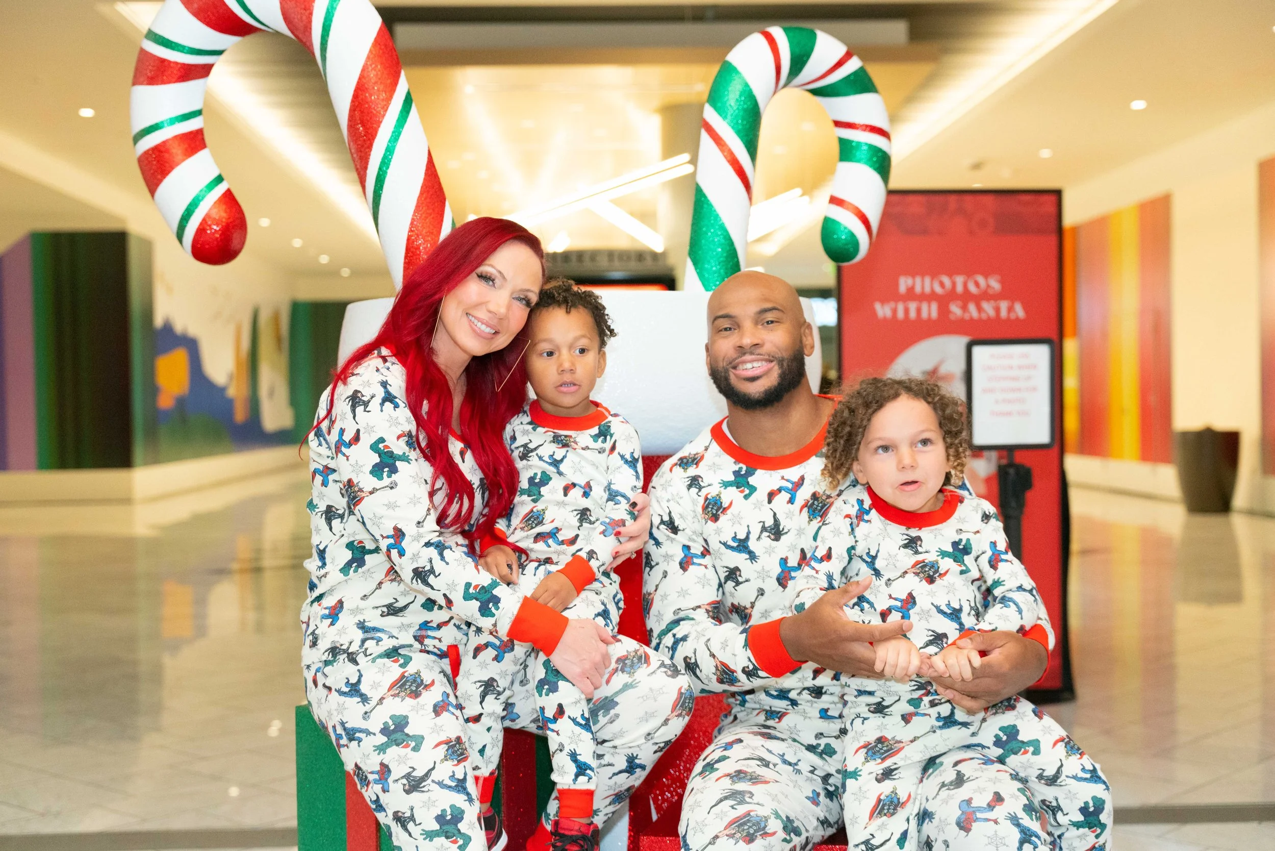 A family of four in Christmas pajamas posing with Santa, with large candy cane decorations and a sign that reads 'Photos With Santa' in a mall.