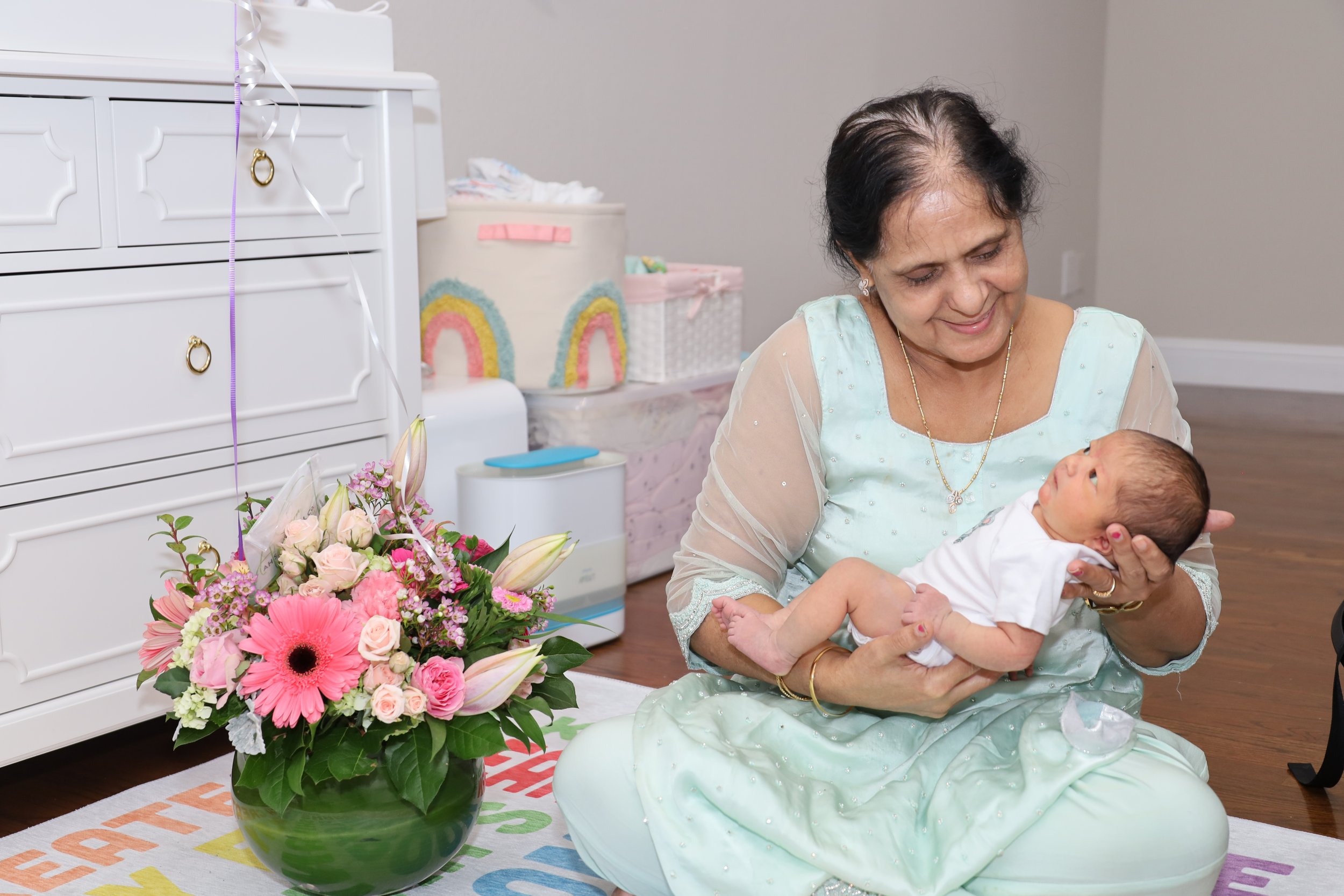 An older woman holding a baby girl in a light green dress, smiling and looking at her. A colorful bouquet of flowers on the floor nearby, with decorations and storage bins in the background.