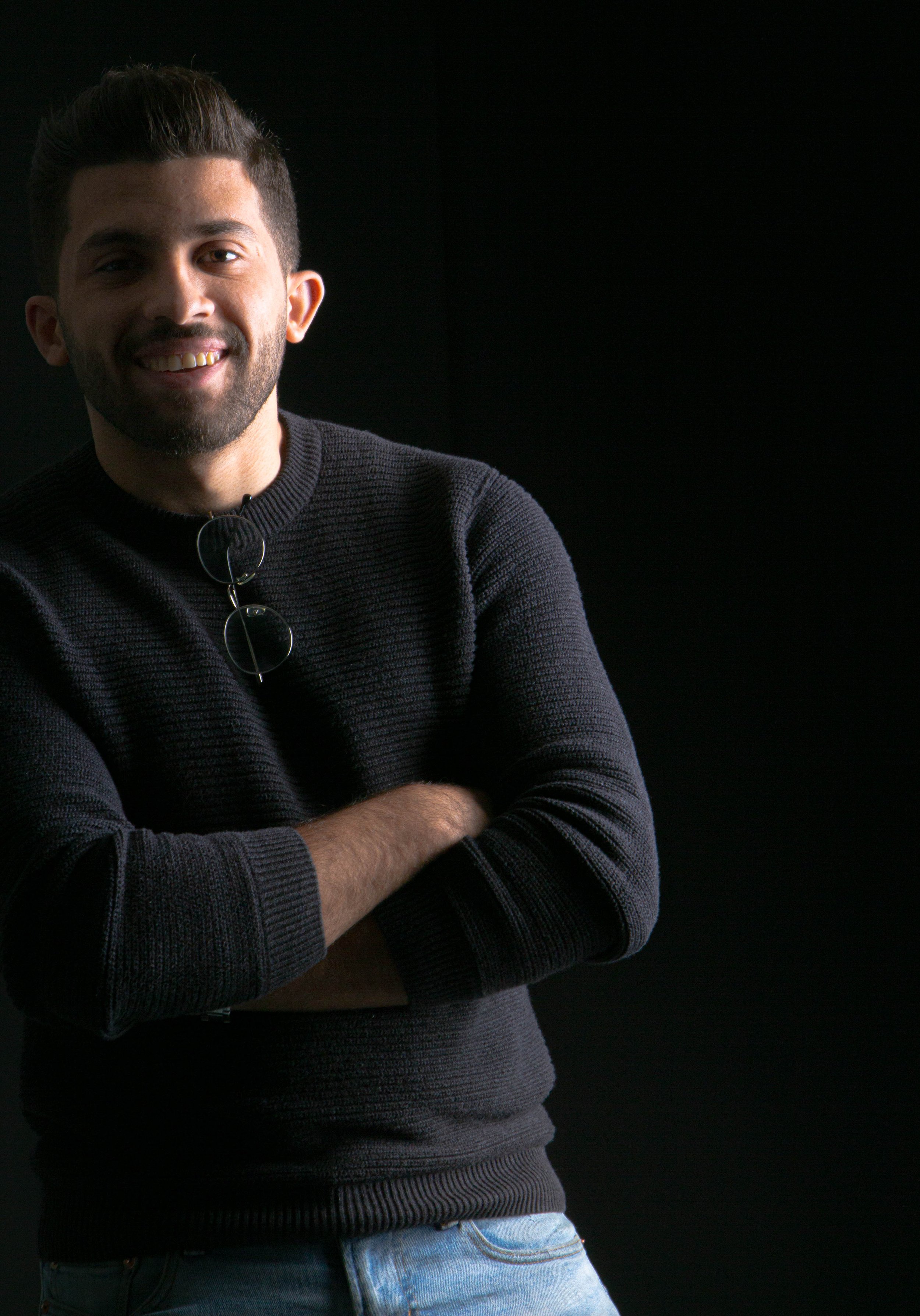 Young man with dark hair and beard smiling, crossing arms, wearing a black sweater and light blue jeans, standing against a dark background.