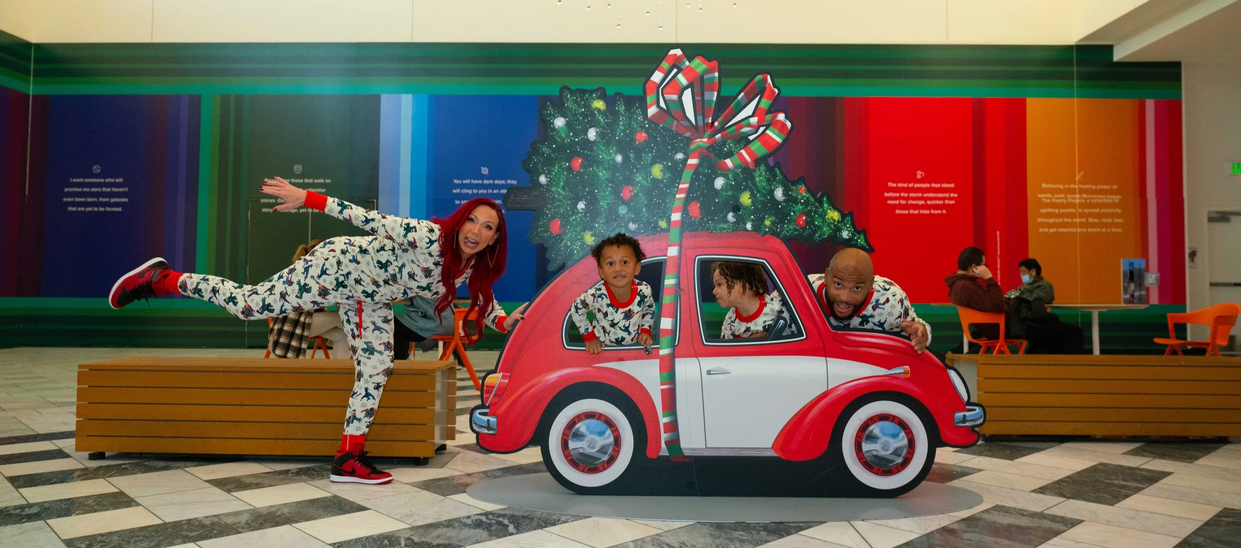 A family dressed in matching Christmas-themed pajamas posing around a cartoon cutout of a red and white car decorated with a large Christmas tree on top, inside a shopping mall with colorful wall art.
