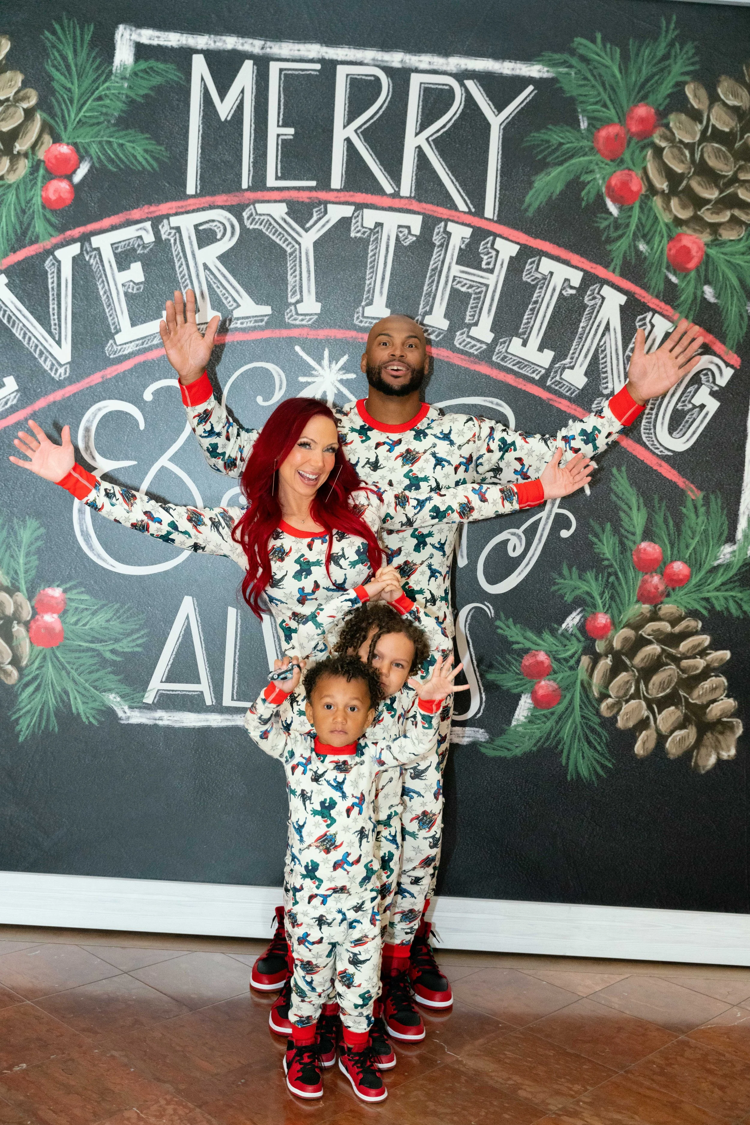 A family of four dressed in matching Christmas pajamas celebrating in front of a festive chalkboard wall with pinecones, holly berries, and the words 'Merry Everything' written on it.