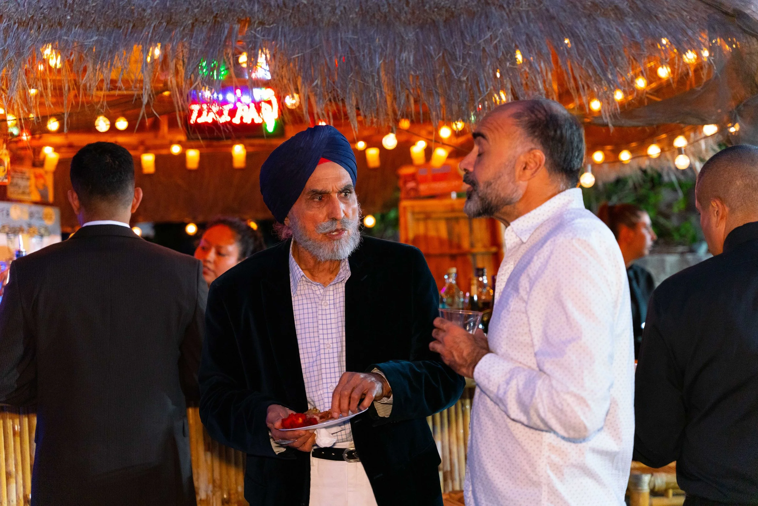 Two men engaged in conversation at a social gathering in a tiki bar or outdoor restaurant, with warm string lights and a thatched roof in the background.