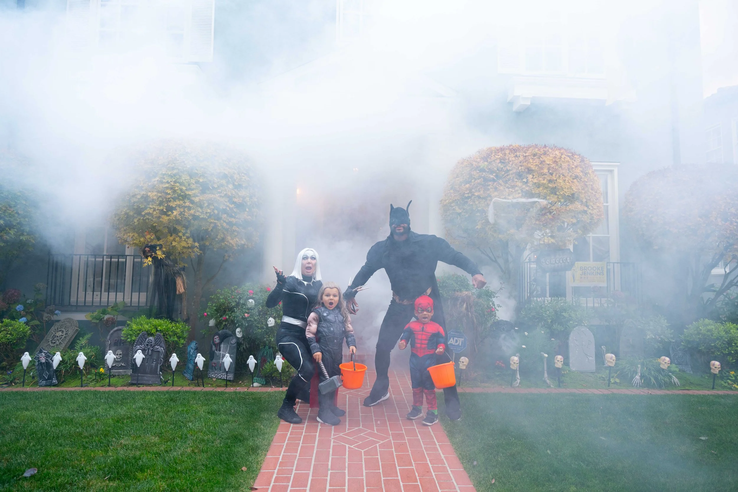 Family dressed in Halloween costumes standing on brick walkway in front of a haunted house with fog and Halloween decorations, including skulls and ghost figures.