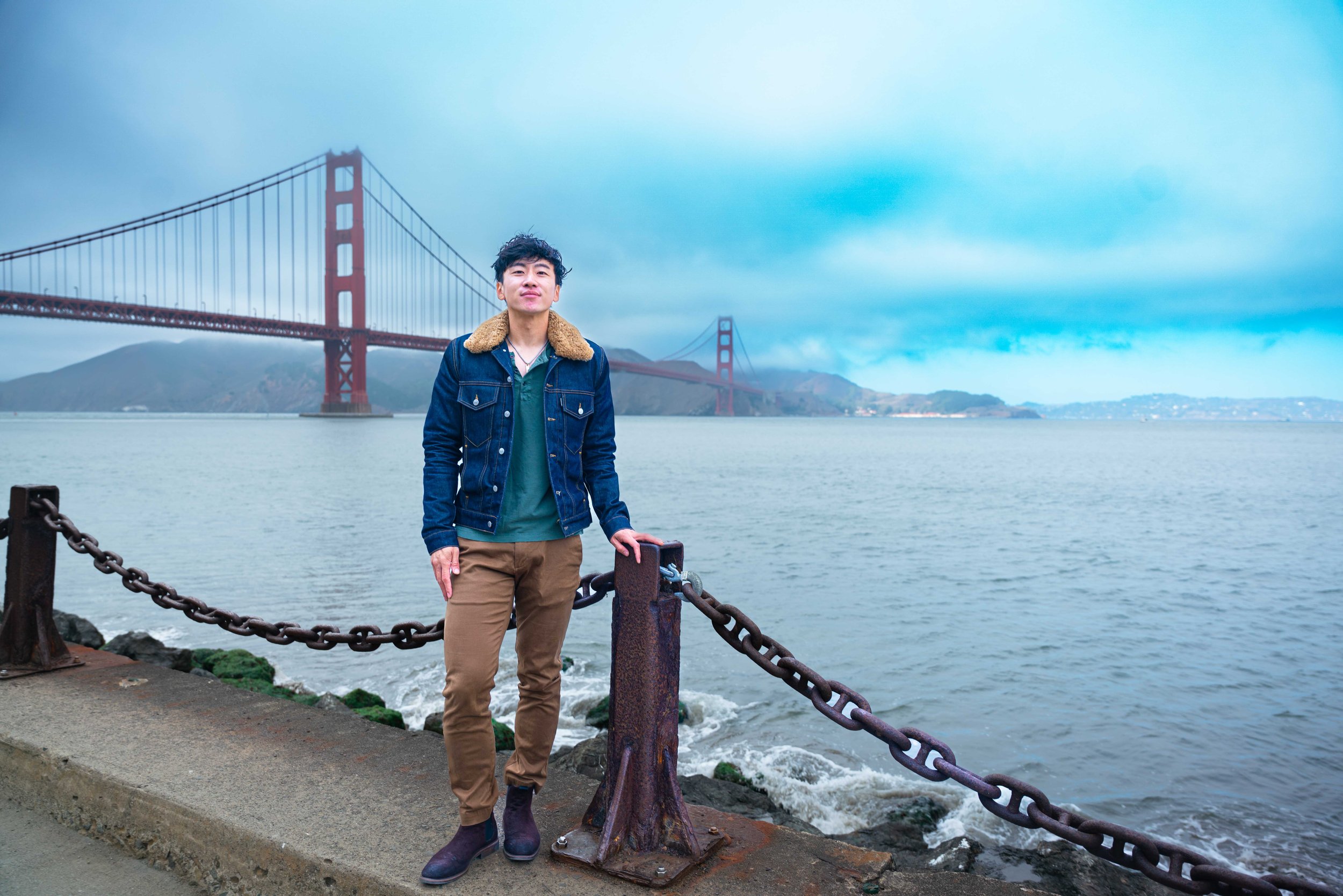 A young man standing by a chain fence near the water with the Golden Gate Bridge in the background, cloudy sky.