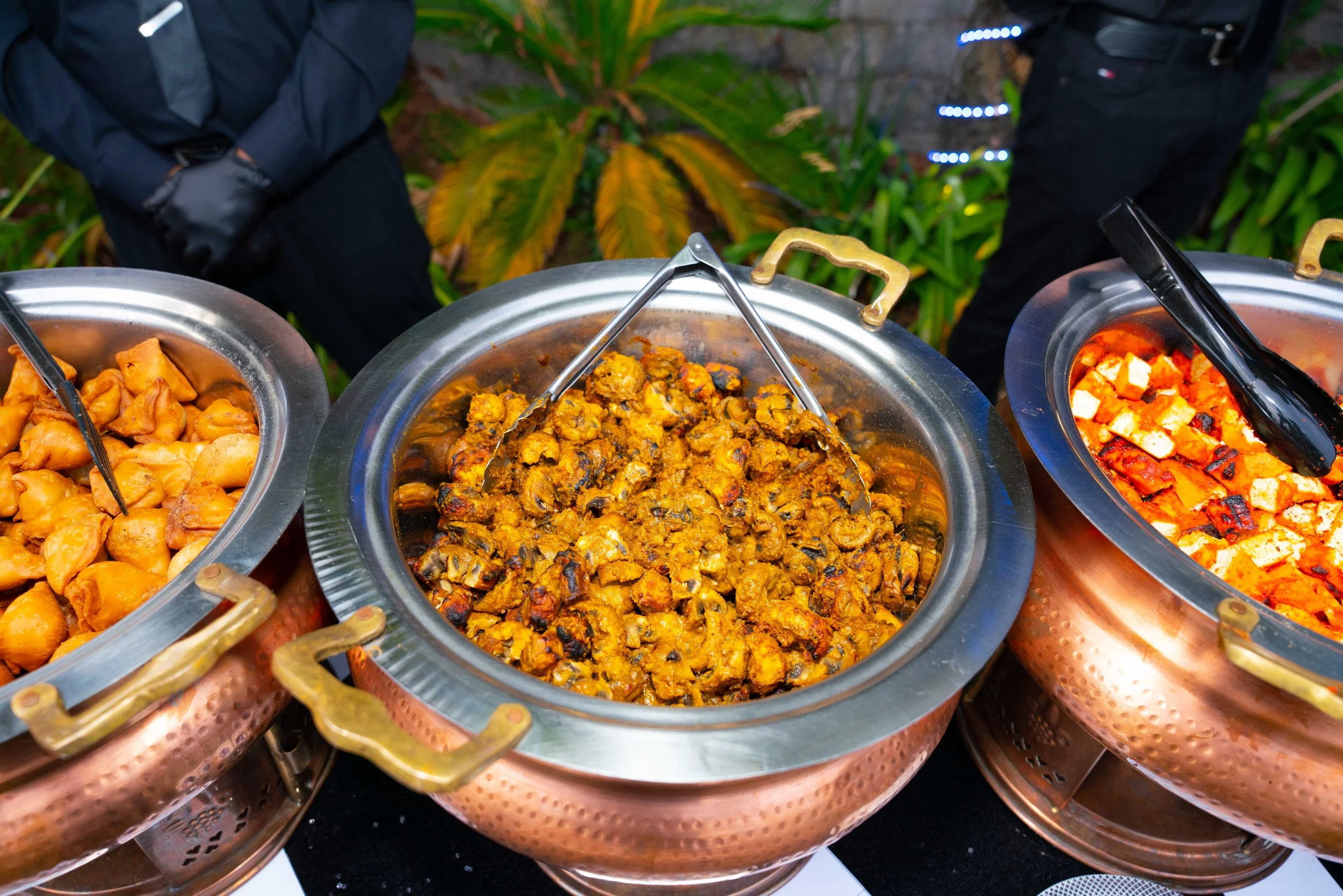 Three large copper serving bowls filled with different types of cooked Indian food, with a background of green plants and partially visible people in dark clothing.