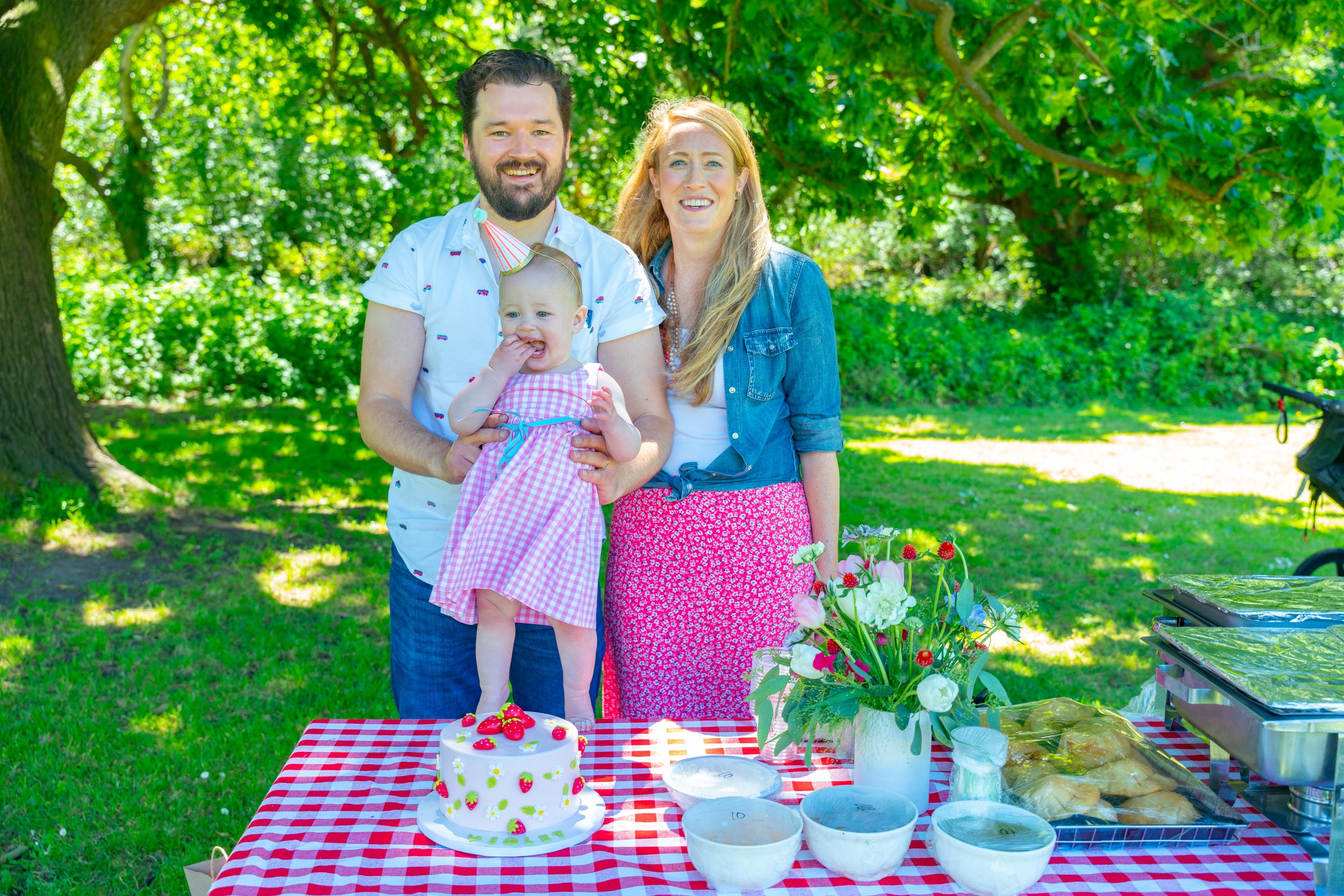Family celebrating outdoors with a birthday cake on a red and white checkered tablecloth. The family includes a man, woman, and a young girl in a pink checkered dress. The man is holding the girl, and they are smiling in a green, leafy park.