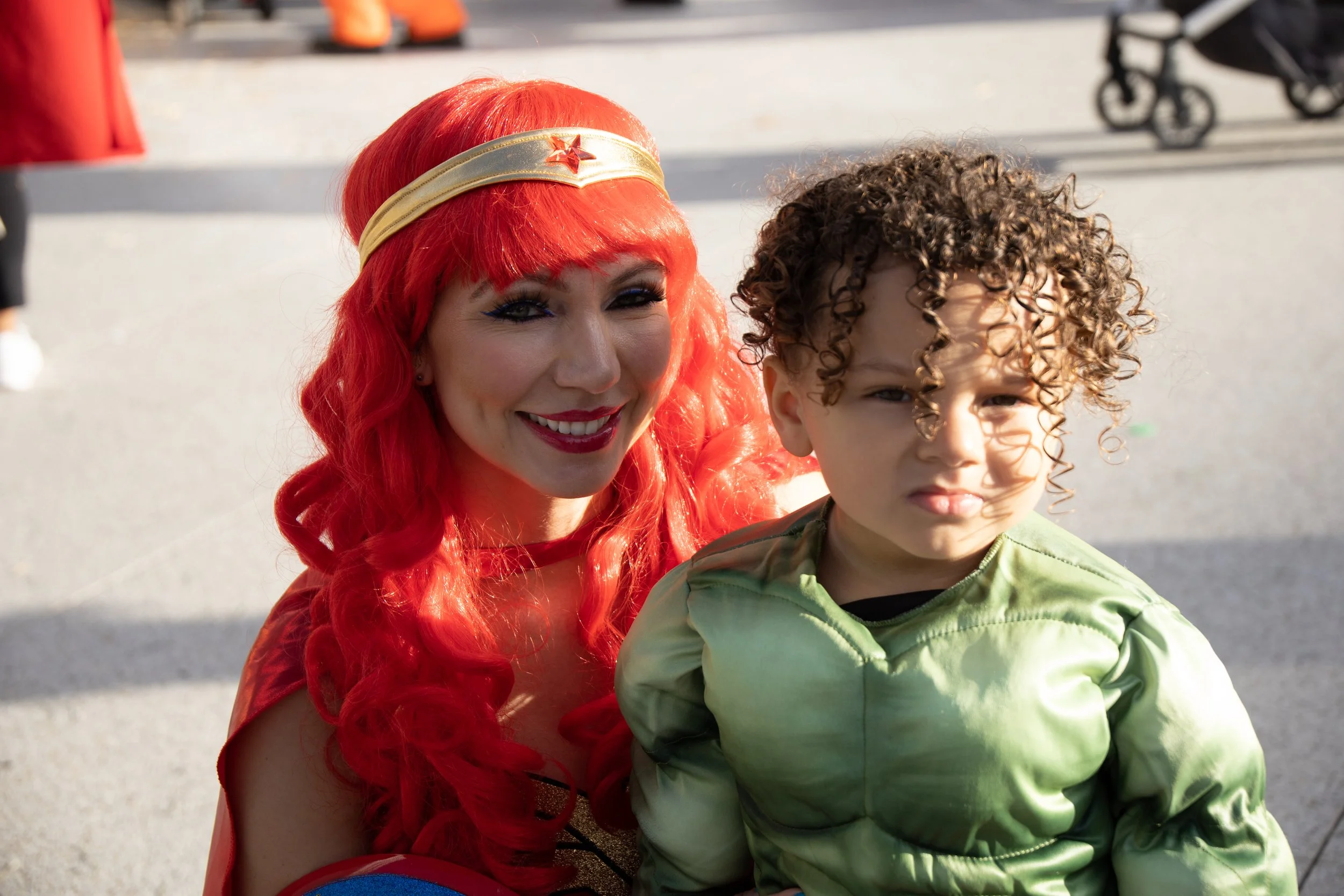 A woman dressed as Wonder Woman with bright red hair and a young boy with curly hair, both outdoors during daytime.