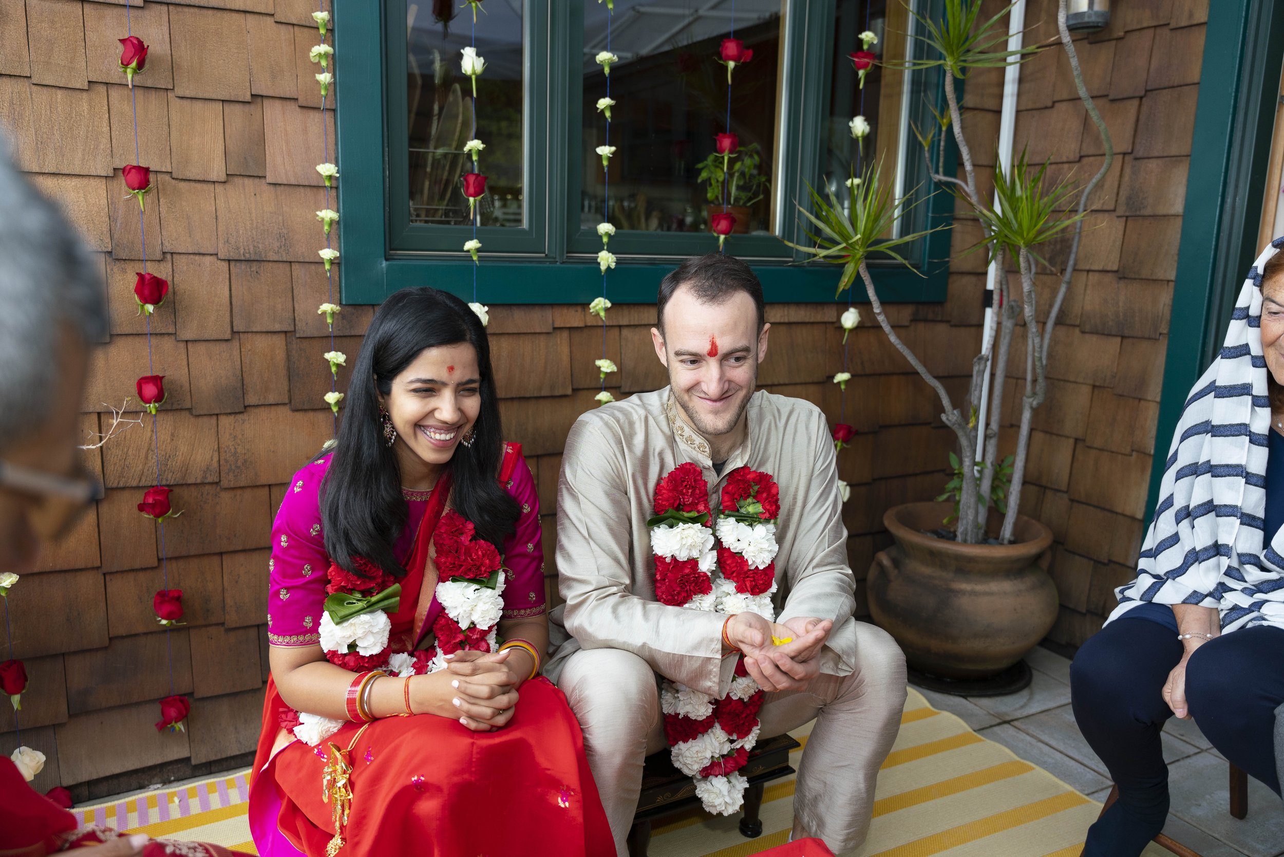 A couple in traditional South Asian wedding attire getting married, seated on a porch decorated with red and white flower garlands, smiling happily.