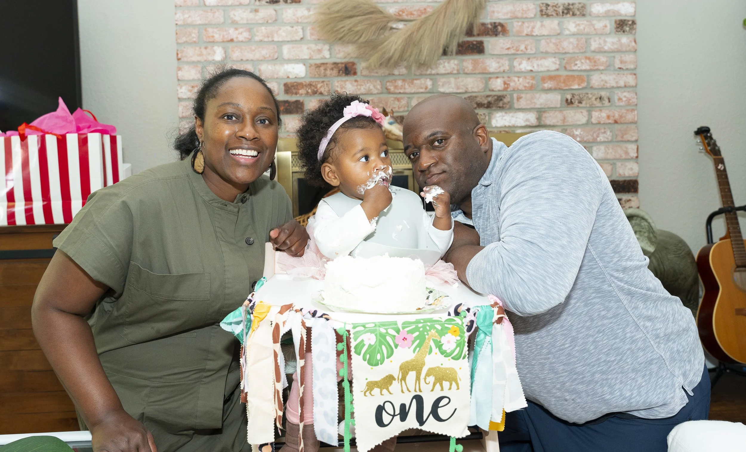 A young girl celebrating her first birthday with her parents, surrounded by a decorated cake and a festive banner, in a cozy indoor setting with a brick wall background.