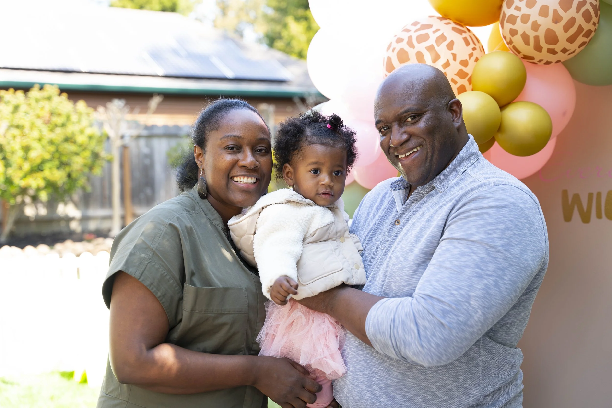 A happy African American family of three, with a woman and a man holding a young girl, smiling outdoors in front of pink and orange balloons at a celebration.