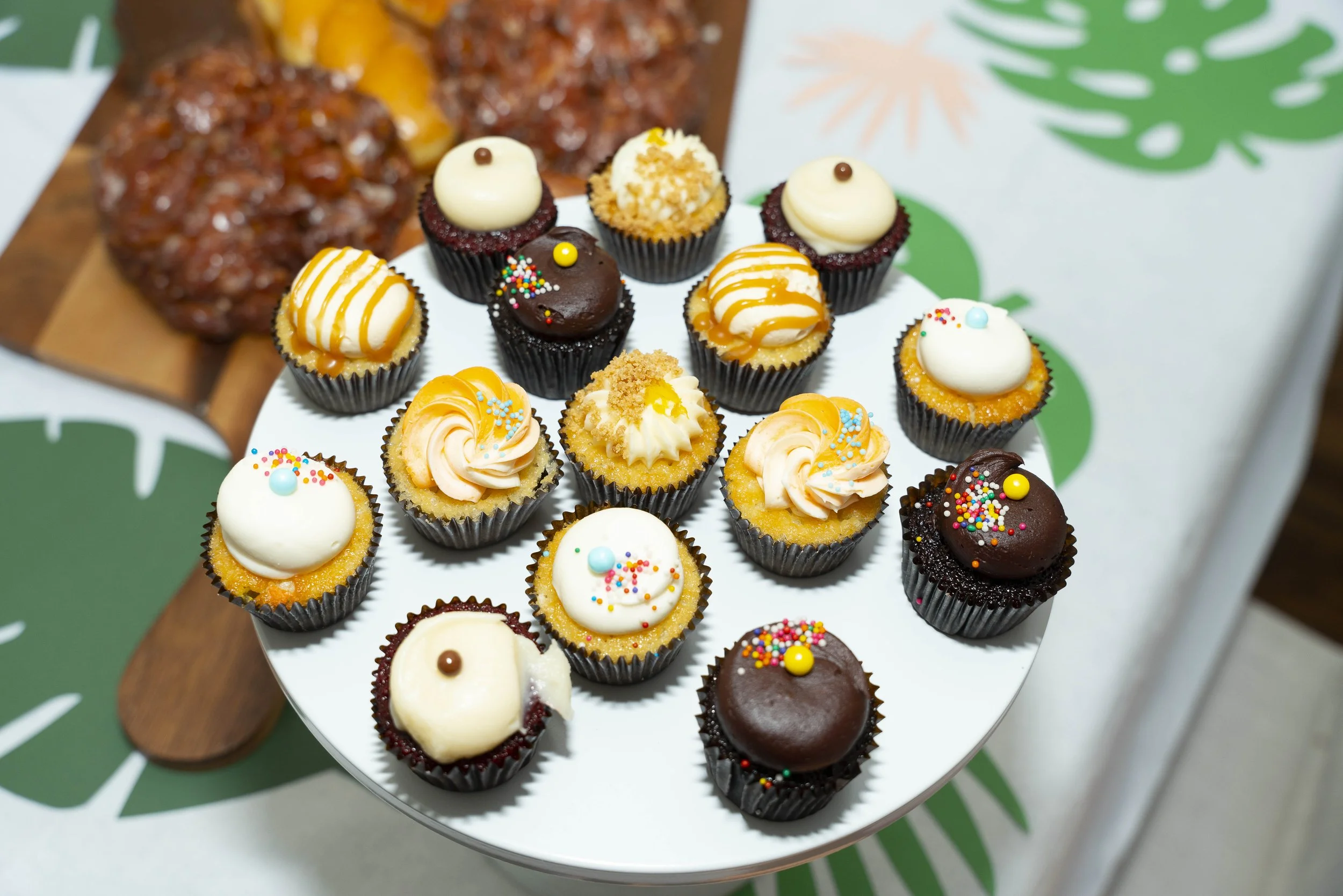 Assorted decorated cupcakes on a white round tray, with some chocolate chip cookies visible in the background.