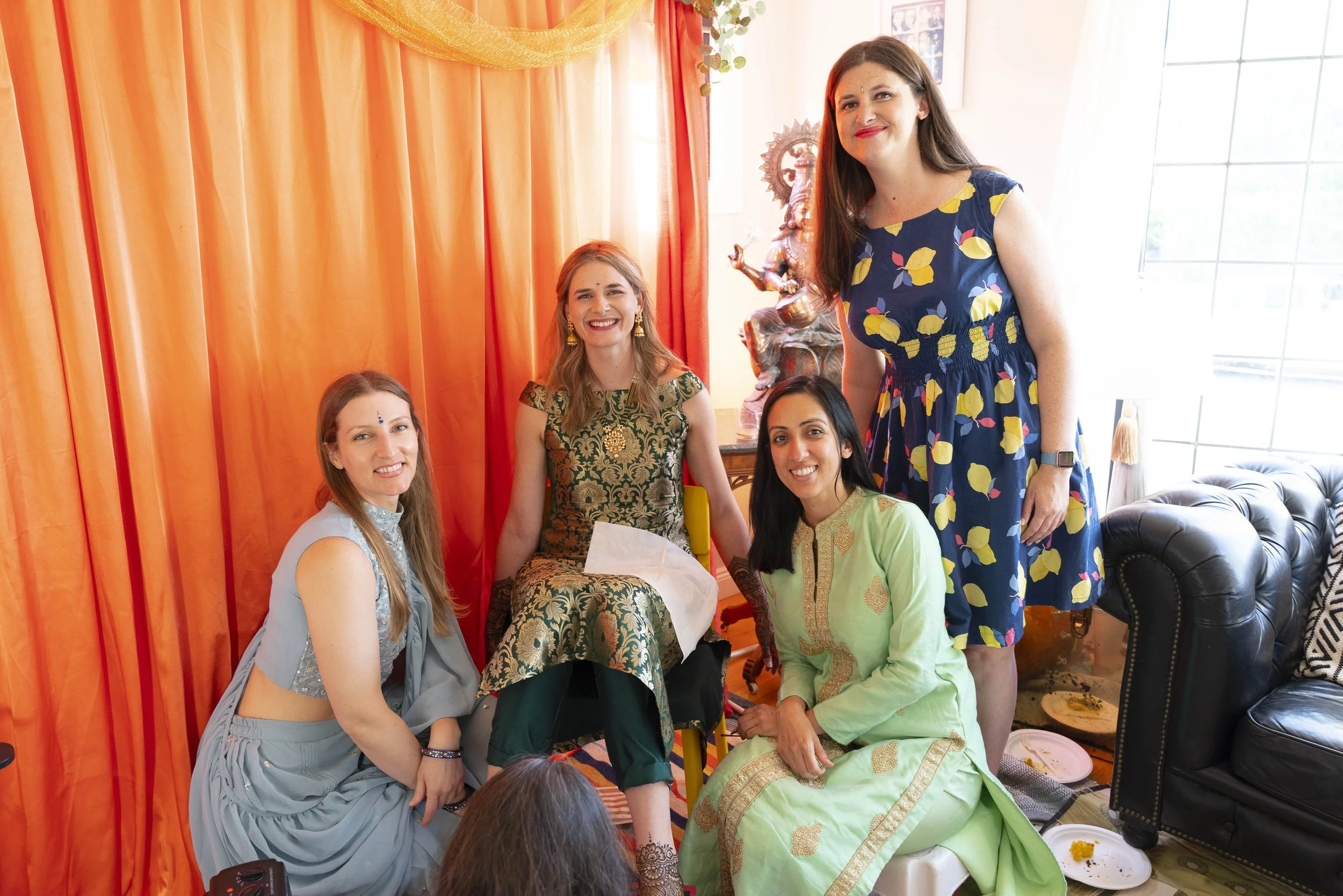 Group of five women celebrating, with bright orange curtains and decorations in the background, one woman sitting in a wheelchair, all smiling and dressed in colorful festive attire.