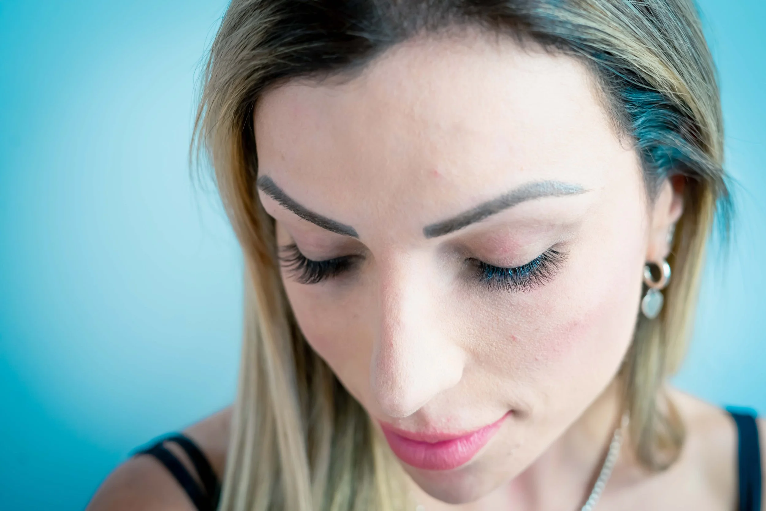 A woman with blonde hair, closed eyes, long eyelashes, and makeup, photographed against a light blue background.