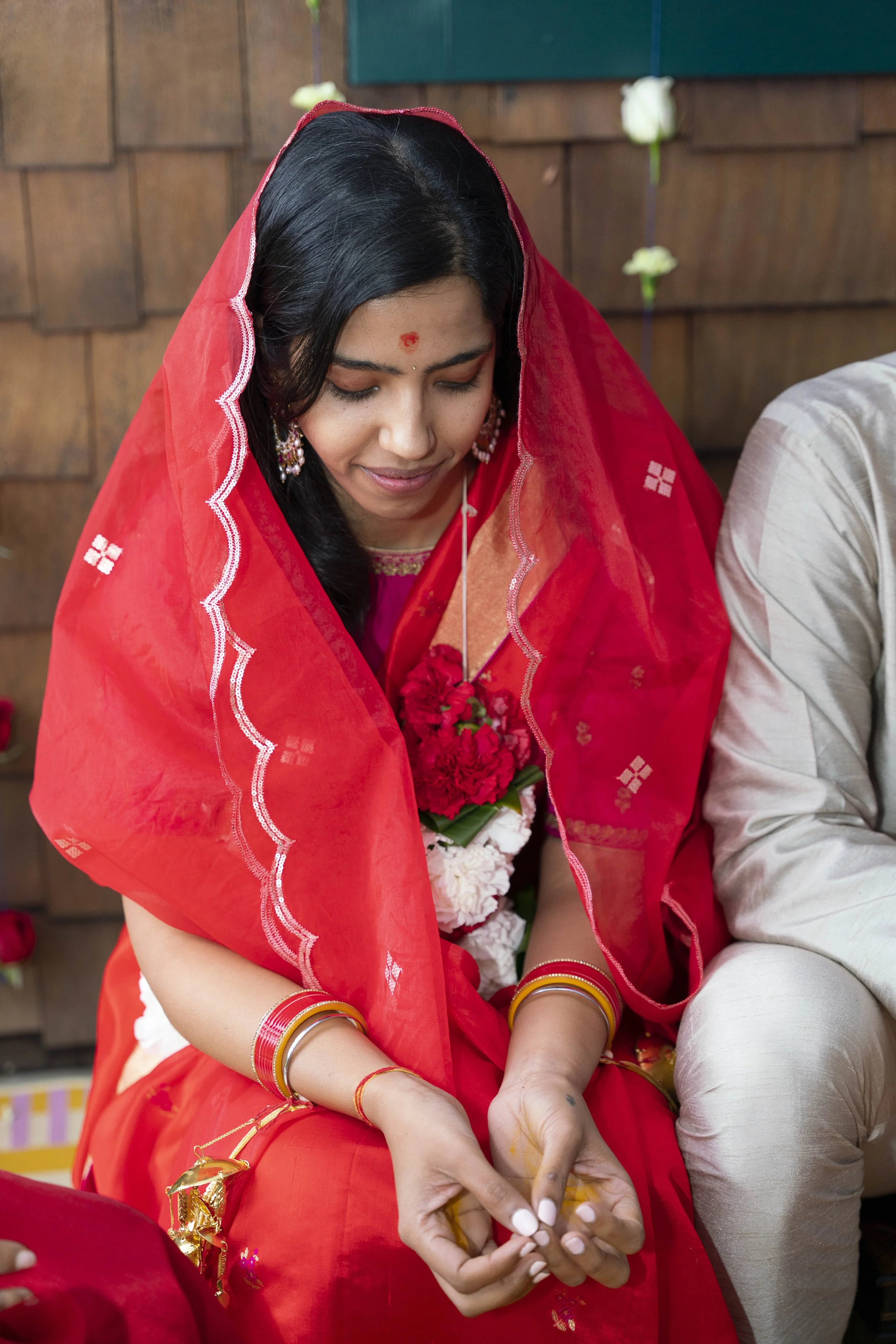 A woman dressed in traditional Indian attire, wearing a red saree with a red veil, sitting with a man during a wedding ceremony, with garlands around her neck and jewelry.