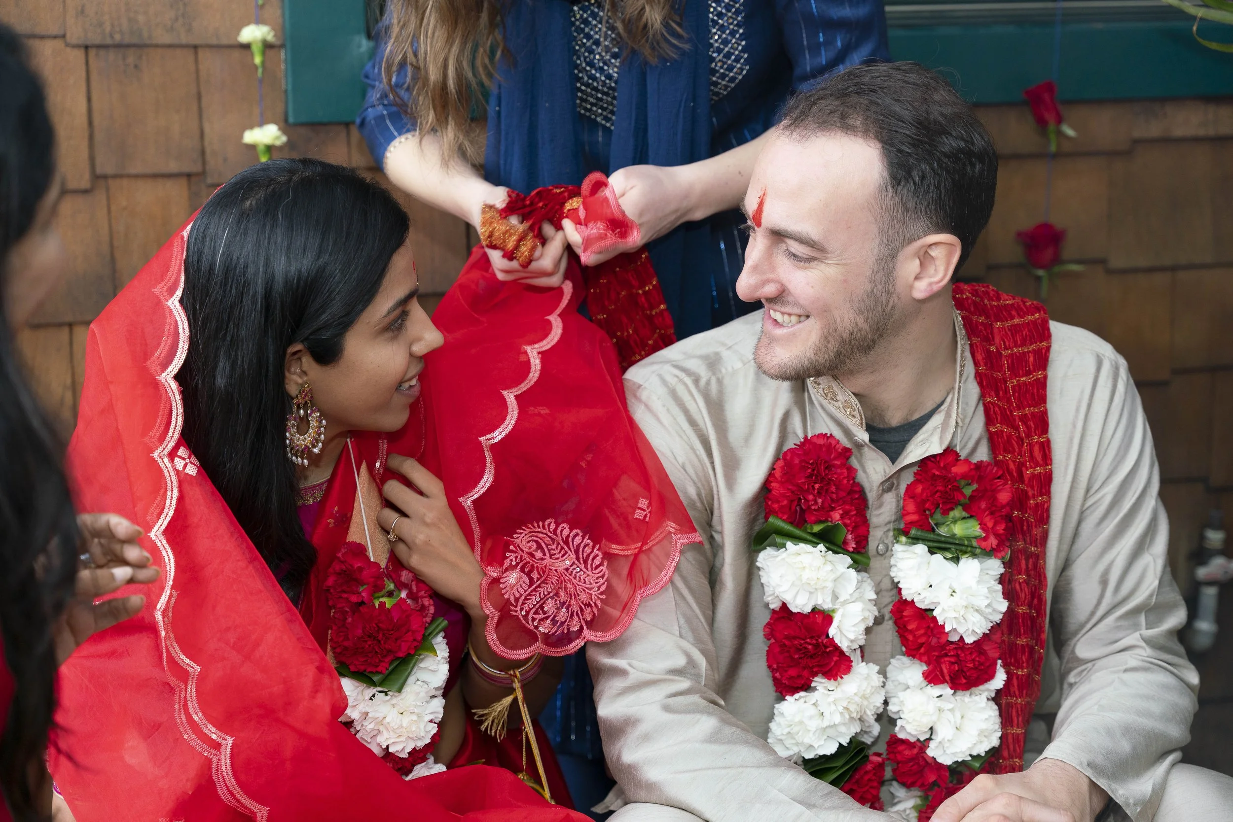A multicultural couple at a wedding ceremony, dressed in traditional Indian attire, exchanging smiles. The bride wears a red sari with intricate embroidery, jewelry, and a red veil over her head. The groom wears a cream-colored kurta with red and whi