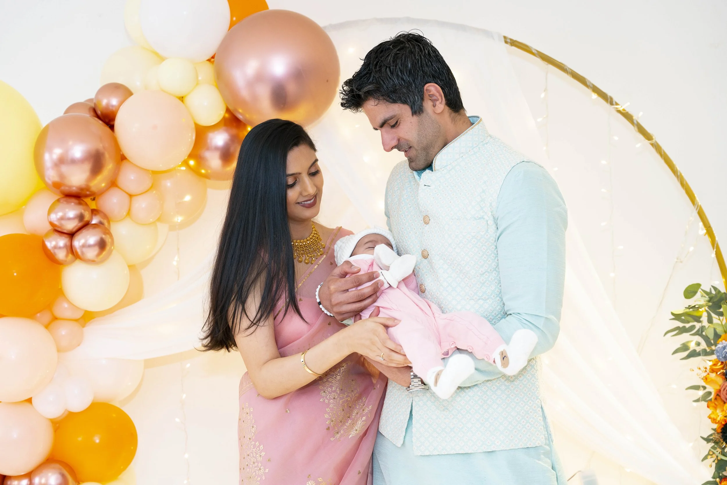 A couple holding a newborn baby during a celebration with balloons and decorative lights.