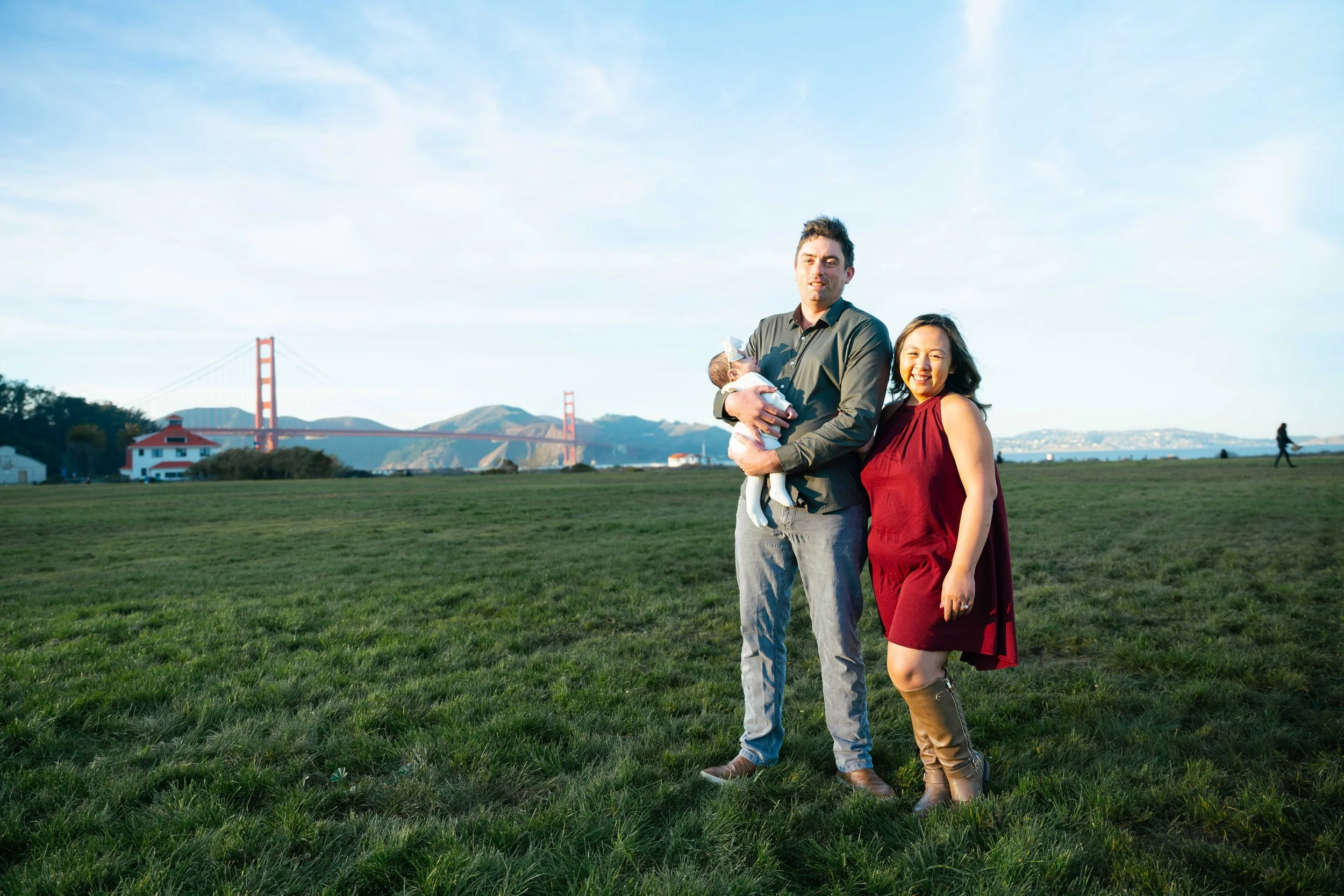 A family of three standing on a grassy field near the Golden Gate Bridge. The father is holding a newborn baby, the mother is smiling and wearing a red dress and brown boots. The background shows a blue sky, a few distant people, and iconic bridge.