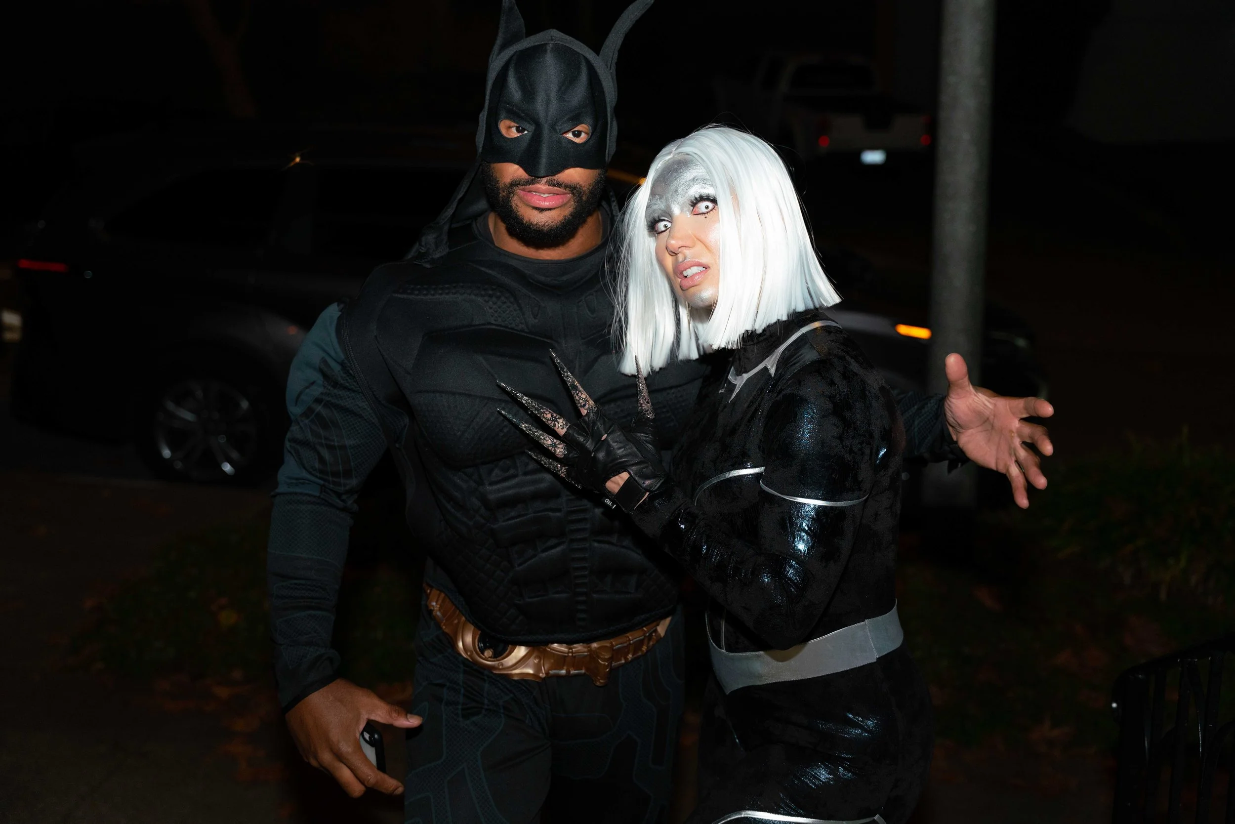 Two people dressed in superhero and villain costumes posing together at night.