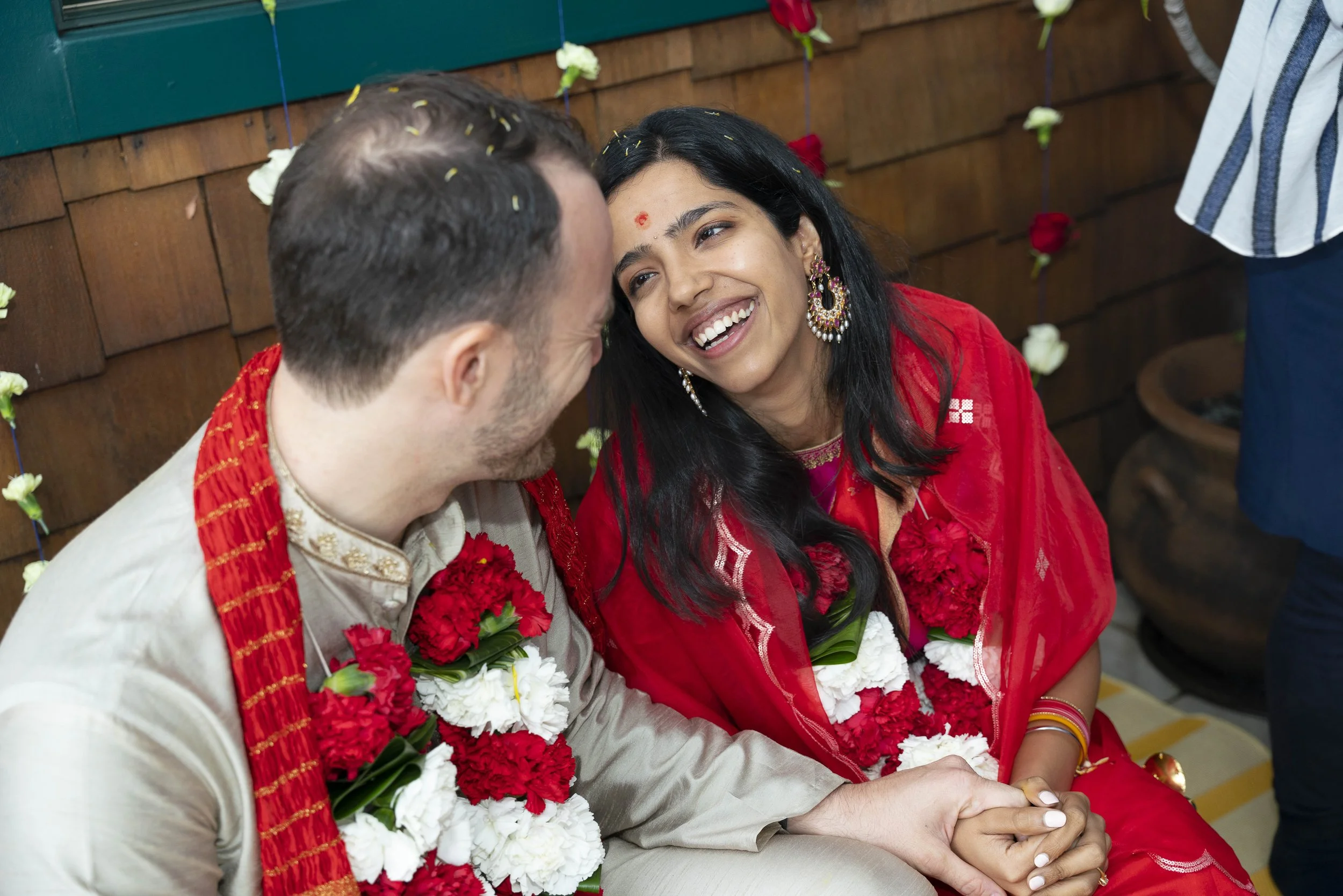 A smiling couple during a wedding or cultural ceremony, wearing traditional attire and floral garlands, sitting close together.
