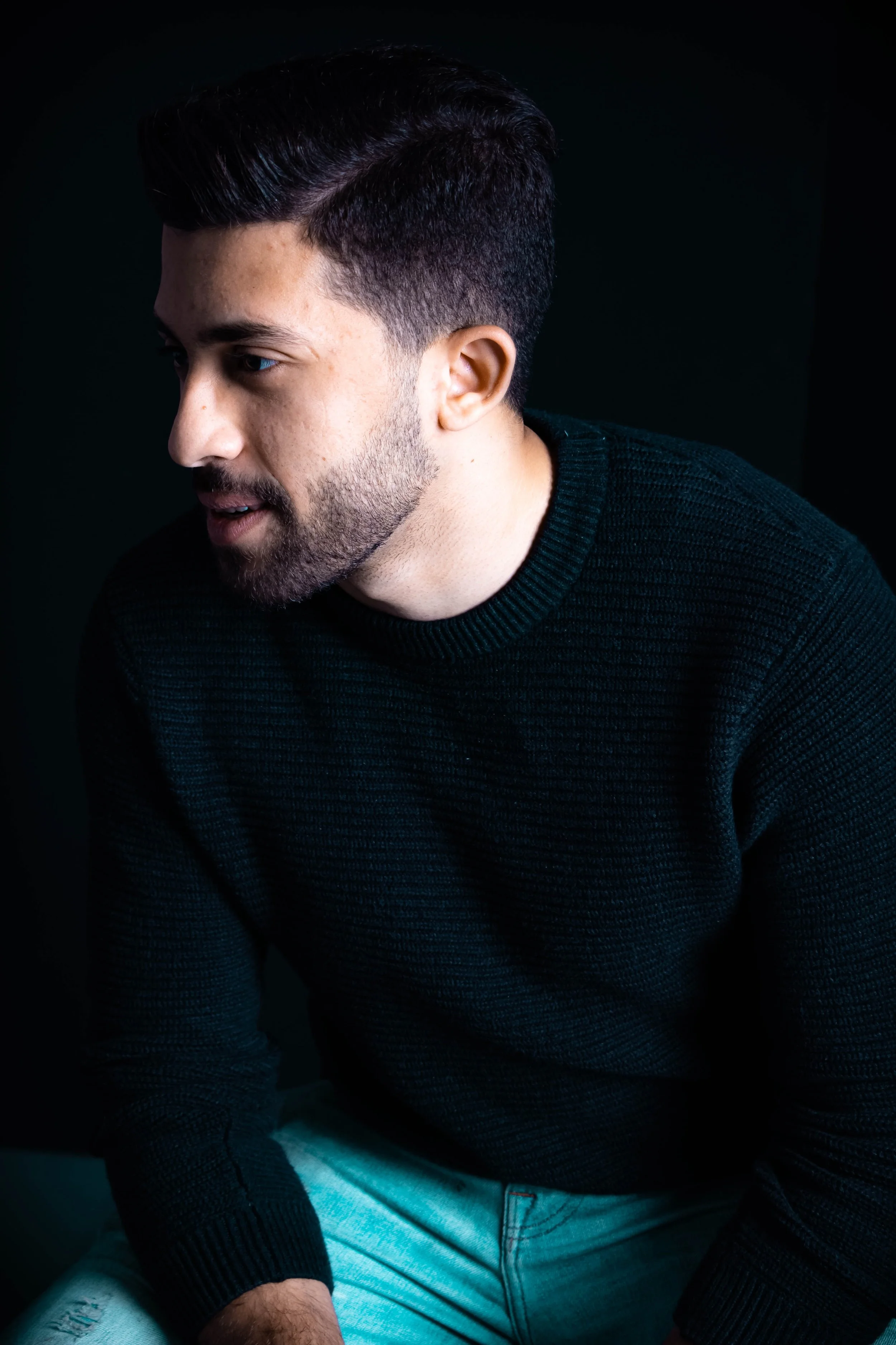 Profile of a young man with dark hair and trimmed beard wearing a black sweater against a dark background.