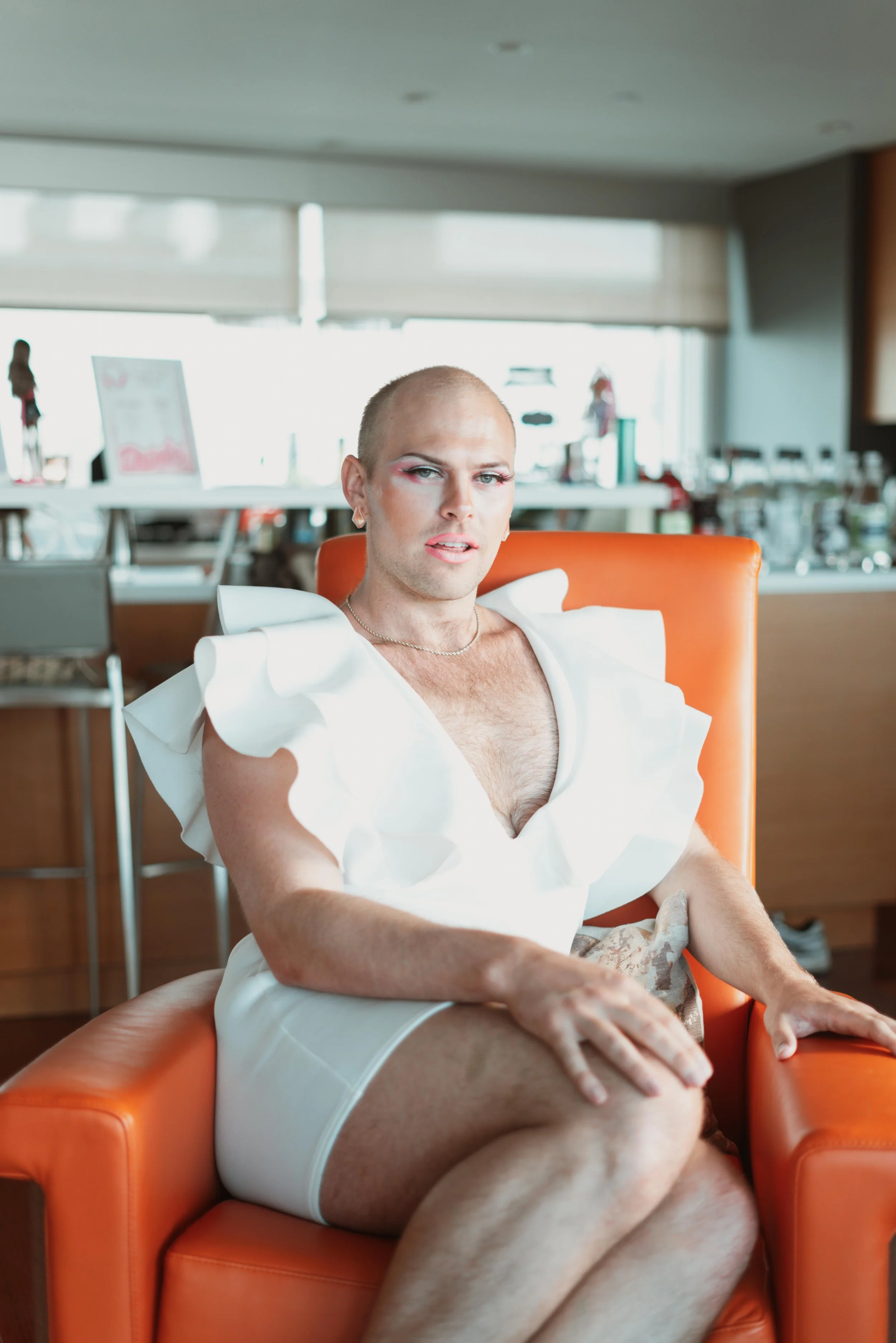 Person sitting in an orange chair in a bright room, wearing a white dress with ruffled sleeves, with a shaved head and makeup.