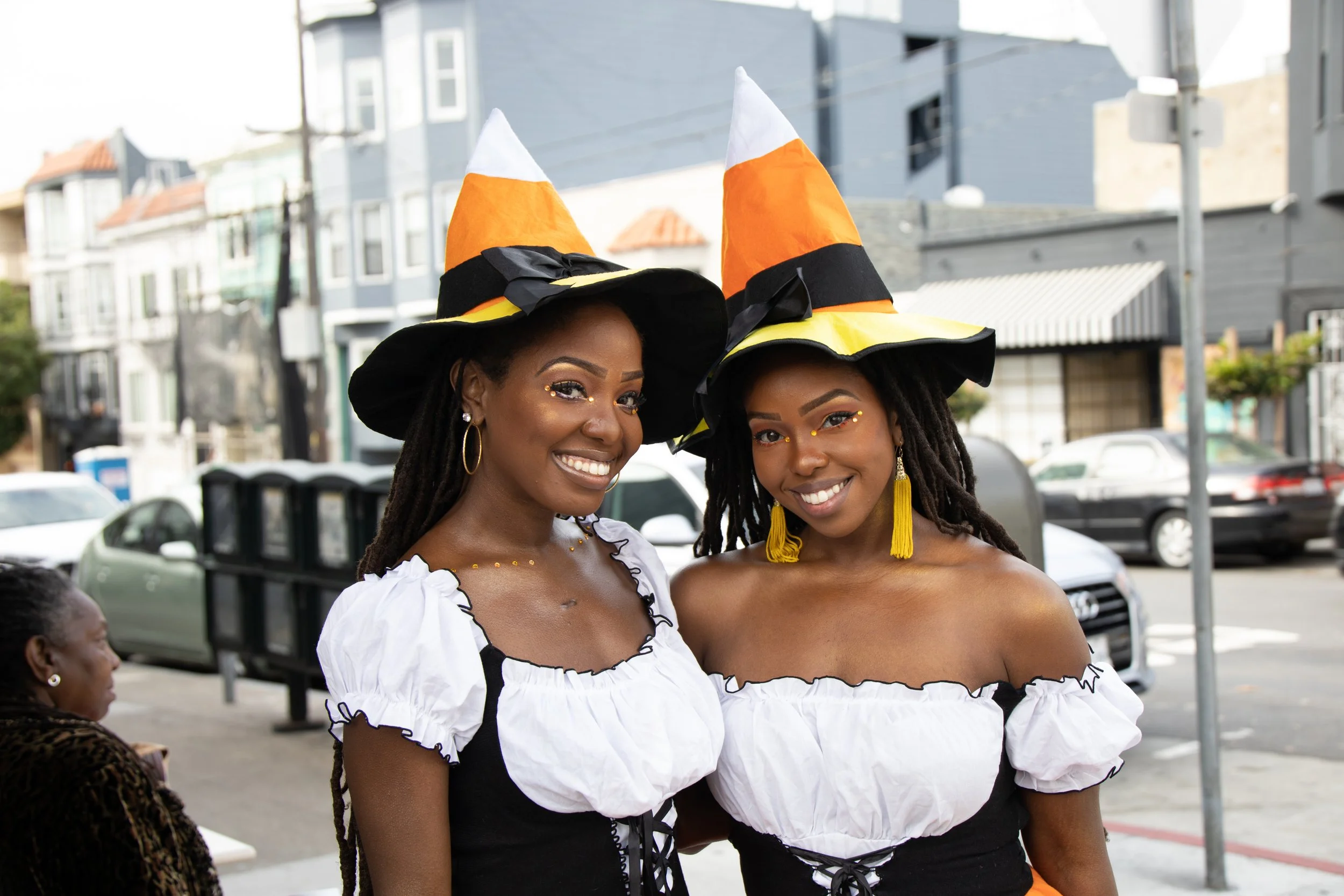 Two women dressed in festive costumes with white off-the-shoulder blouses and black corsets, wearing large Halloween-themed hats with orange, black, and white stripes, smiling and standing outdoors on a street.