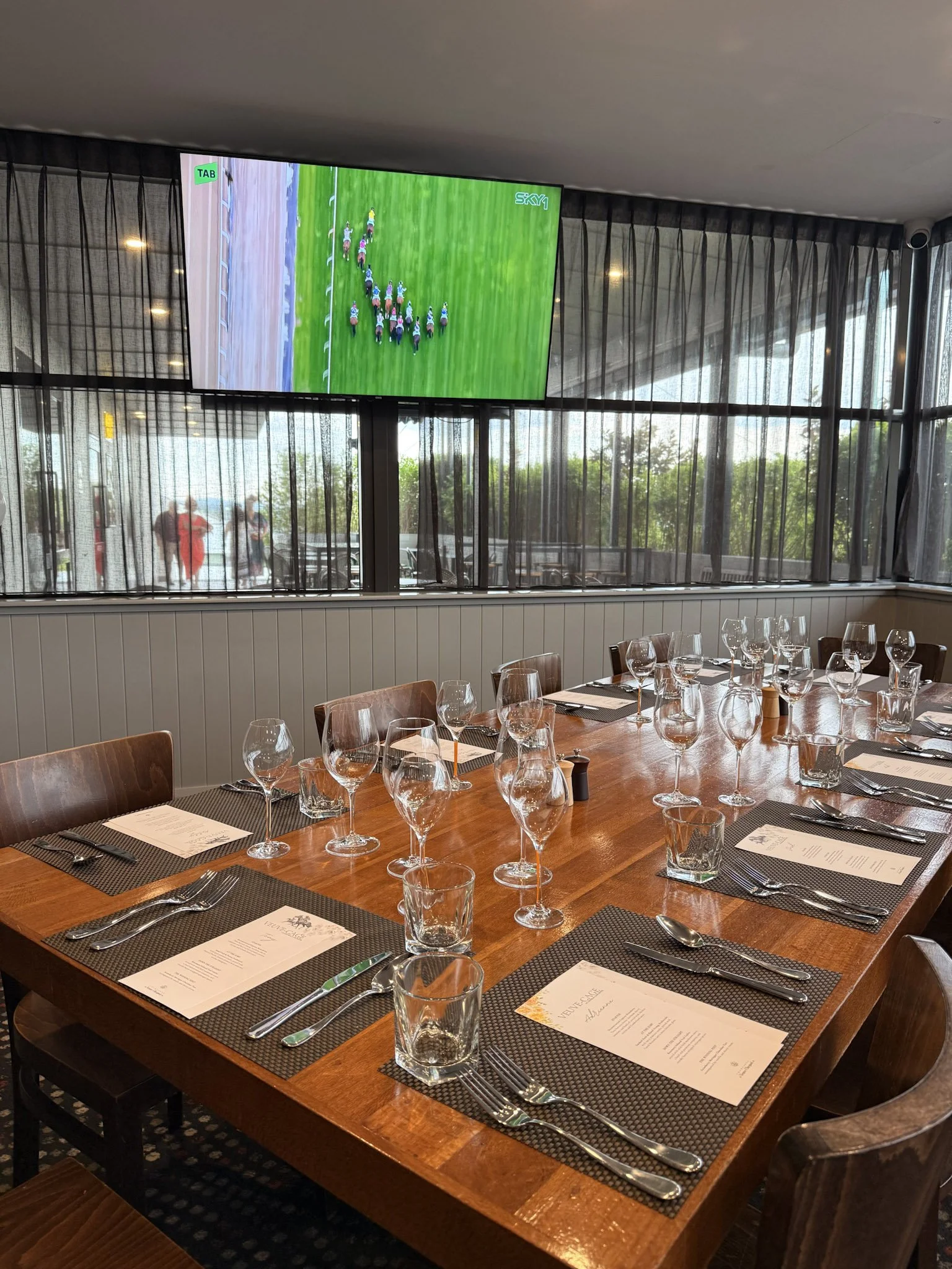 A dining table set with menus, silverware, glasses, and salt and pepper shakers in a restaurant with a large window and a TV showing a horse race.