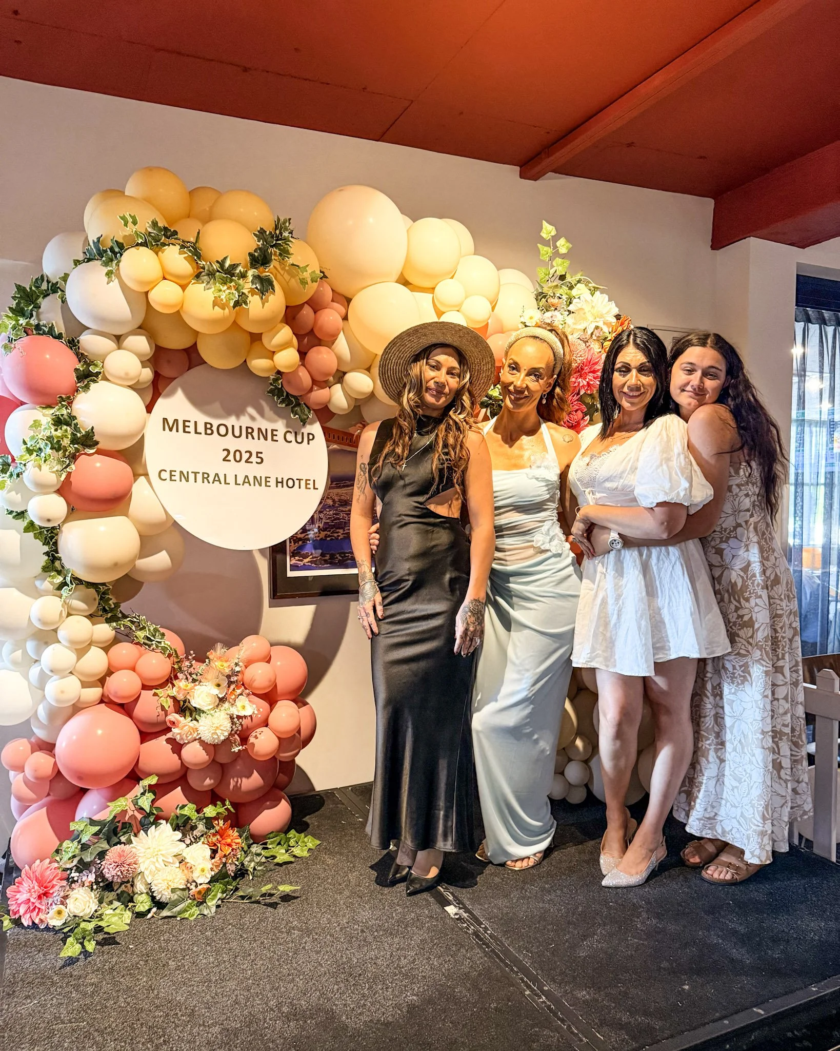 Four women standing together in front of a balloon and flower backdrop, celebrating the Melbourne Cup 2025 at Central Lane Hotel.