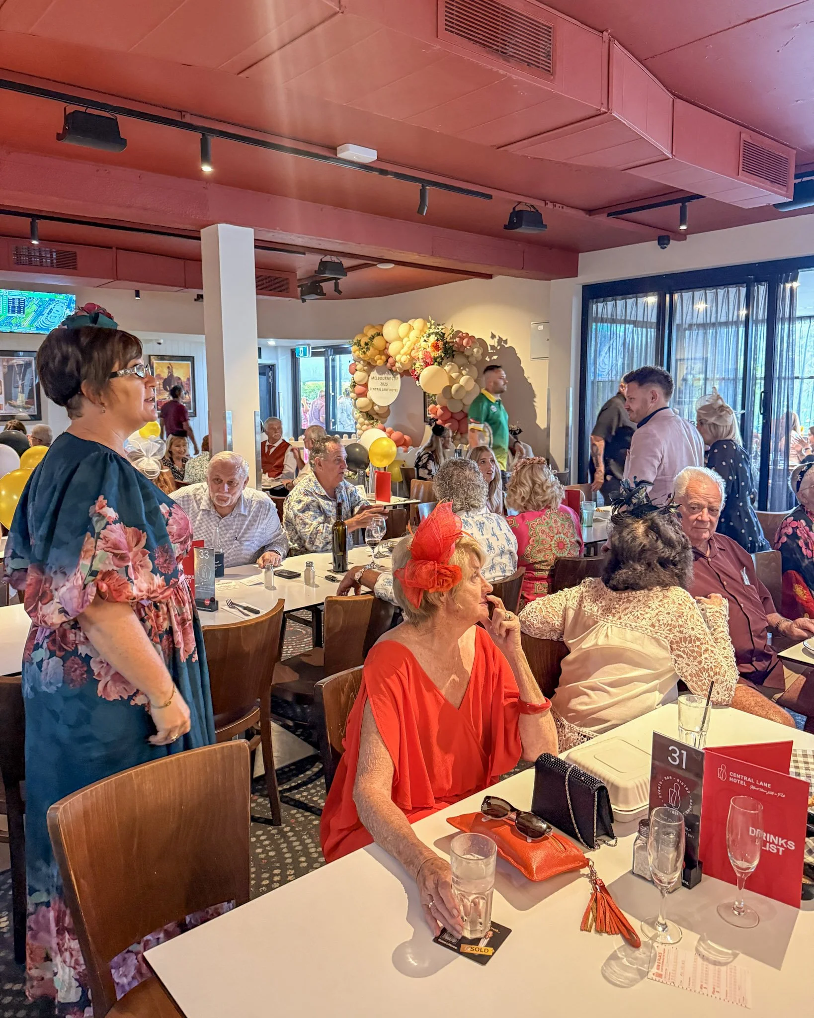 People celebrating at a restaurant decorated with balloons and flowers, with a focus on an elderly woman in a red dress and headpiece sitting at a table.