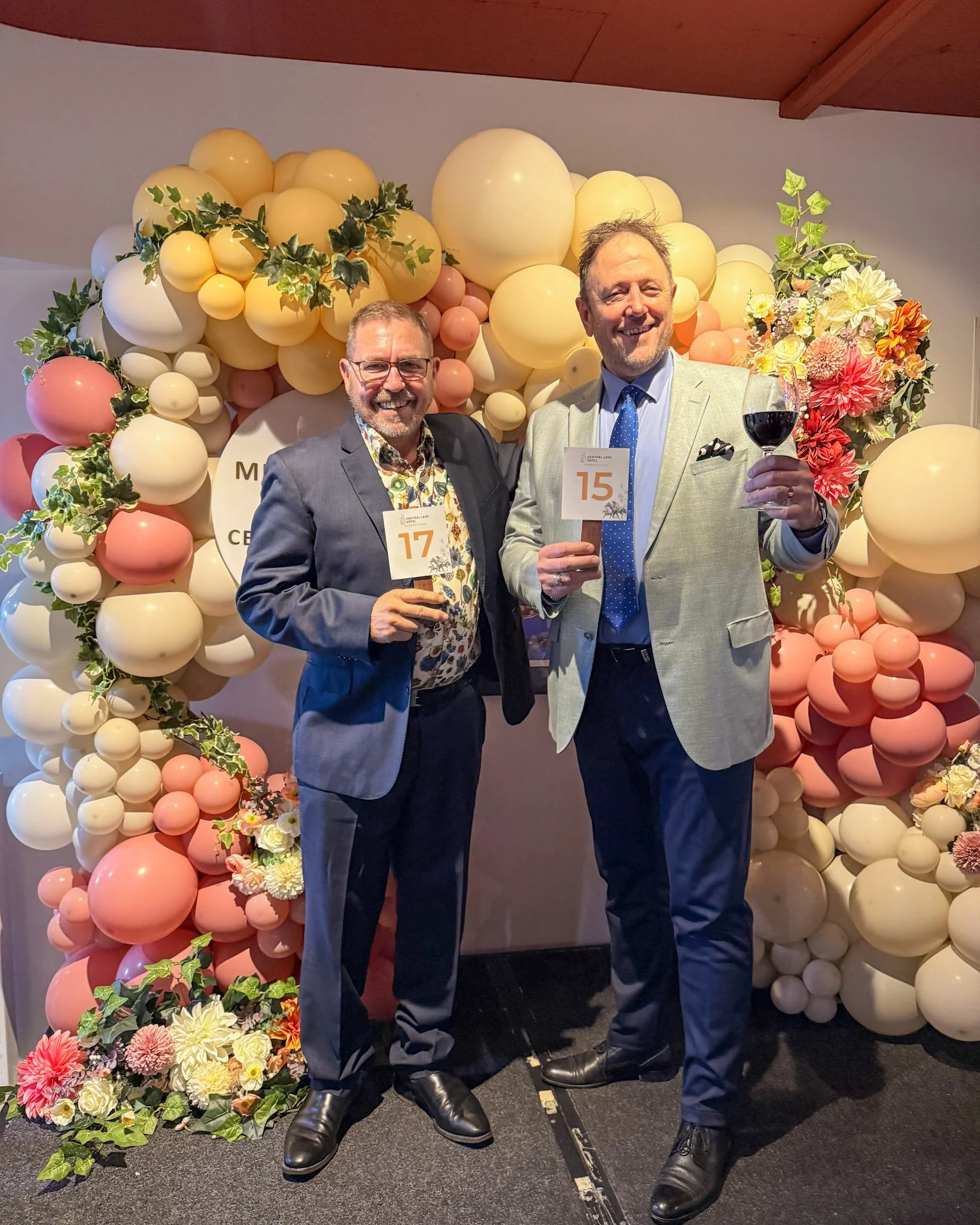 Two men dressed in suits standing in front of a colorful balloon and floral backdrop at a formal event, holding numbered placards and a glass of red wine.