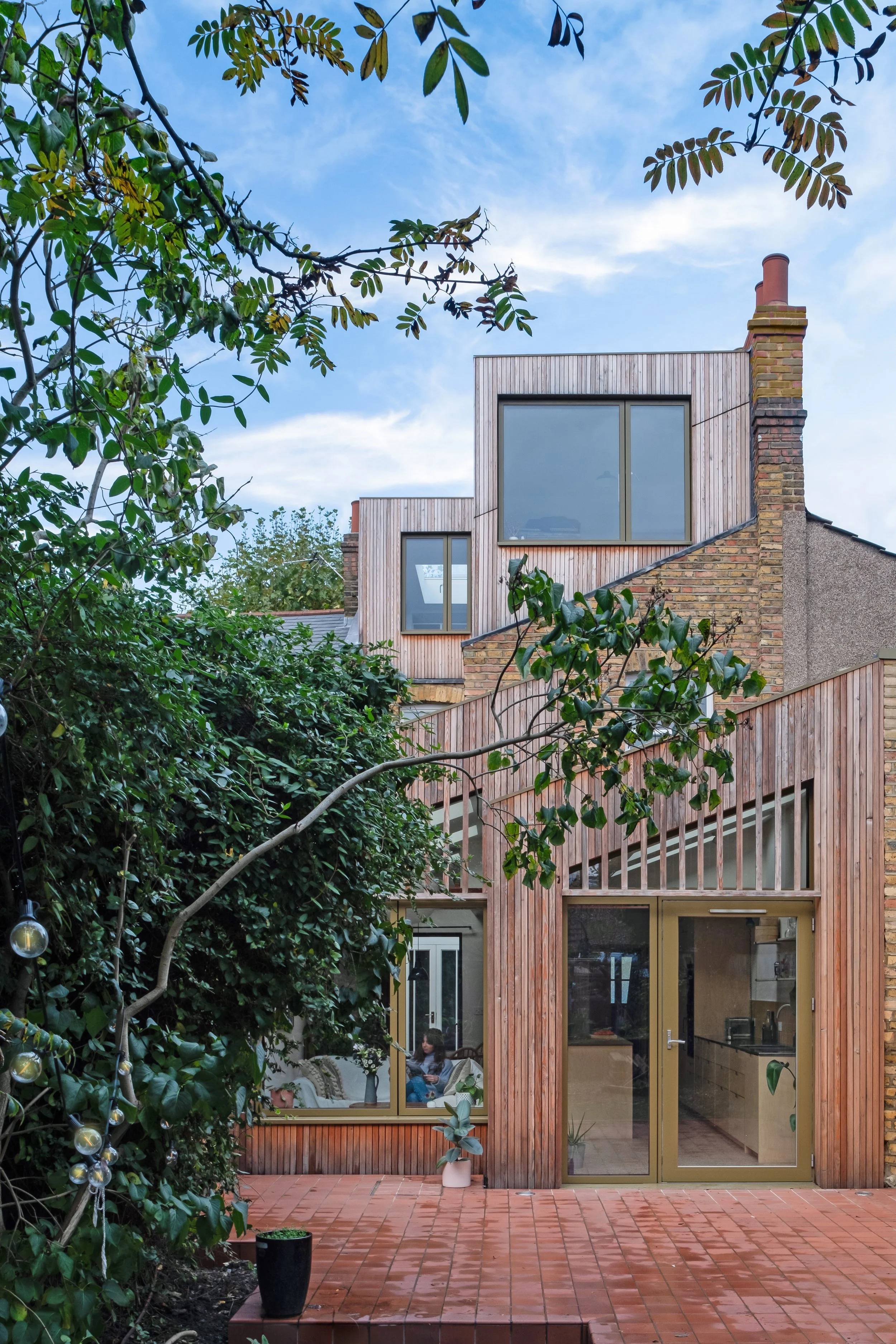 Studio Manifest — Modern house with brick and wooden exterior, large windows, and a red tiled patio, surrounded by greenery and trees, under a blue sky with some clouds