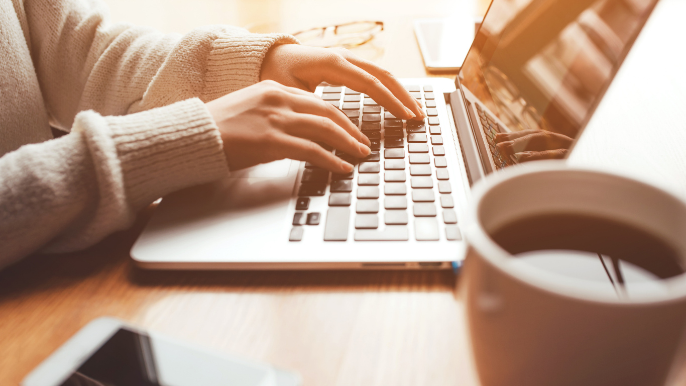 Person tippt auf einer Laptop-Tastatur, eine Tasse Kaffee steht im Vordergrund auf einem Holztisch, in der Nähe liegen ein Smartphone und eine Brille.
