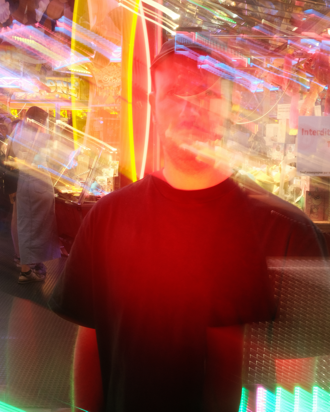 A person in a black shirt standing in front of a brightly lit amusement park attraction with colorful neon lights, city signs, and a blurred figure in the background.
