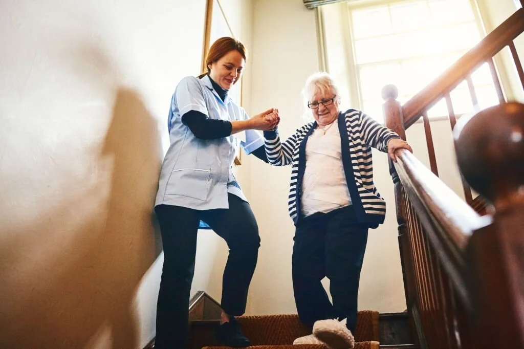 A caregiver helps an elderly woman descend a staircase inside a house, holding her hand for support.