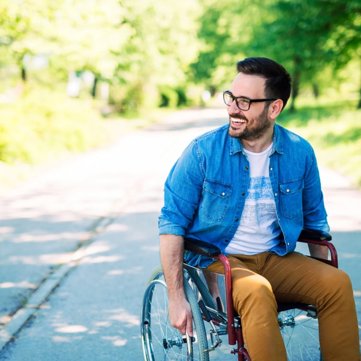 A man with glasses and a beard, wearing a blue jean shirt and brown pants, sitting in a wheelchair outdoors on a sunny day, smiling and looking to his right. The background shows a path with green trees.