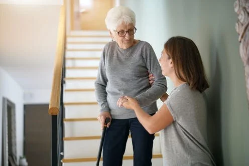 An elderly woman with white hair and glasses holding a cane and standing on stairs, looking at a younger woman seated on the floor, holding her arm, in a home setting.