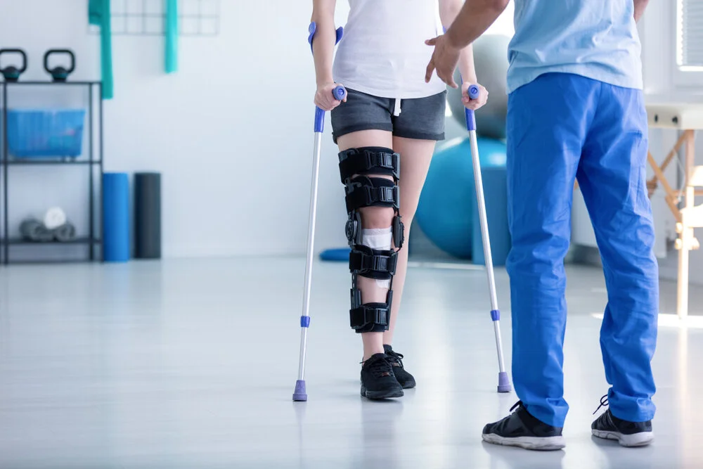 Physical therapist assisting a patient with a knee injury, who is wearing a knee brace and walking with crutches.